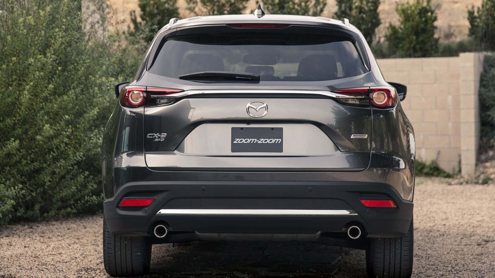 Mazda Cx-9 桌布 壁紙 — 一輛時尚灰色Mazda CX-9 SUV的後視圖，停在碎石上，背景有綠色灌木叢和石牆。