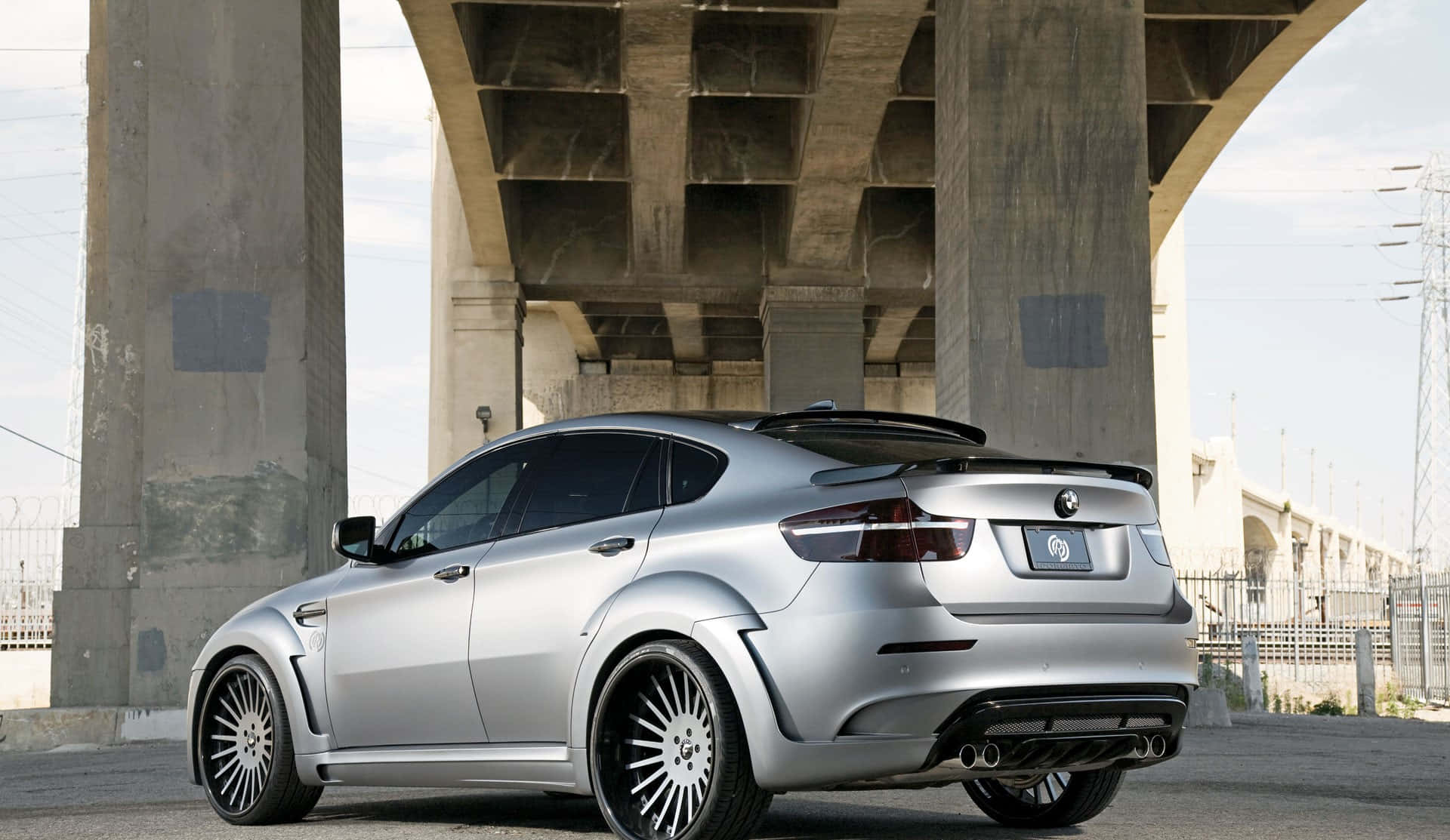 一輛時尚的 BMW X6 M，呈現美麗的金屬灰 壁紙 — 一輛銀色改裝BMW M6雙門跑車停泊在混凝土天橋下，背景有工業圍欄和電線。