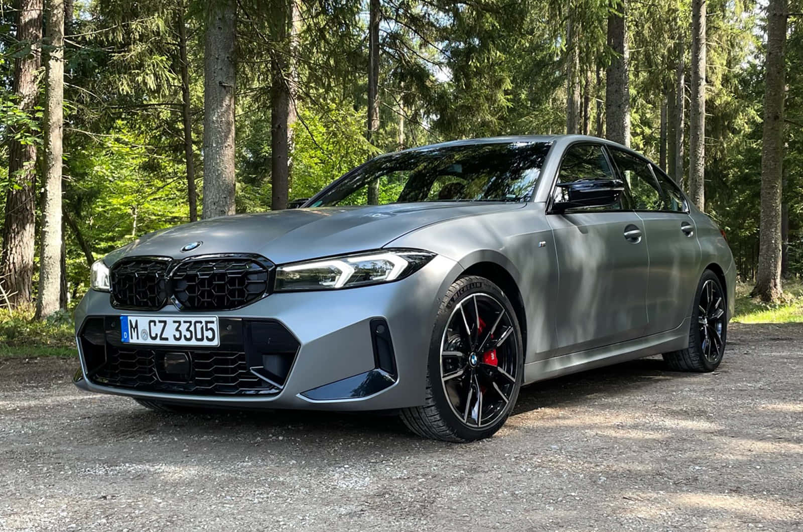 Bmw 3 Series 桌布 壁紙 — 一輛時尚的啞光灰色BMW M3轎車，配備黑色輪圈和紅色制動卡鉗，停在陽光普照的松樹林碎石地上。