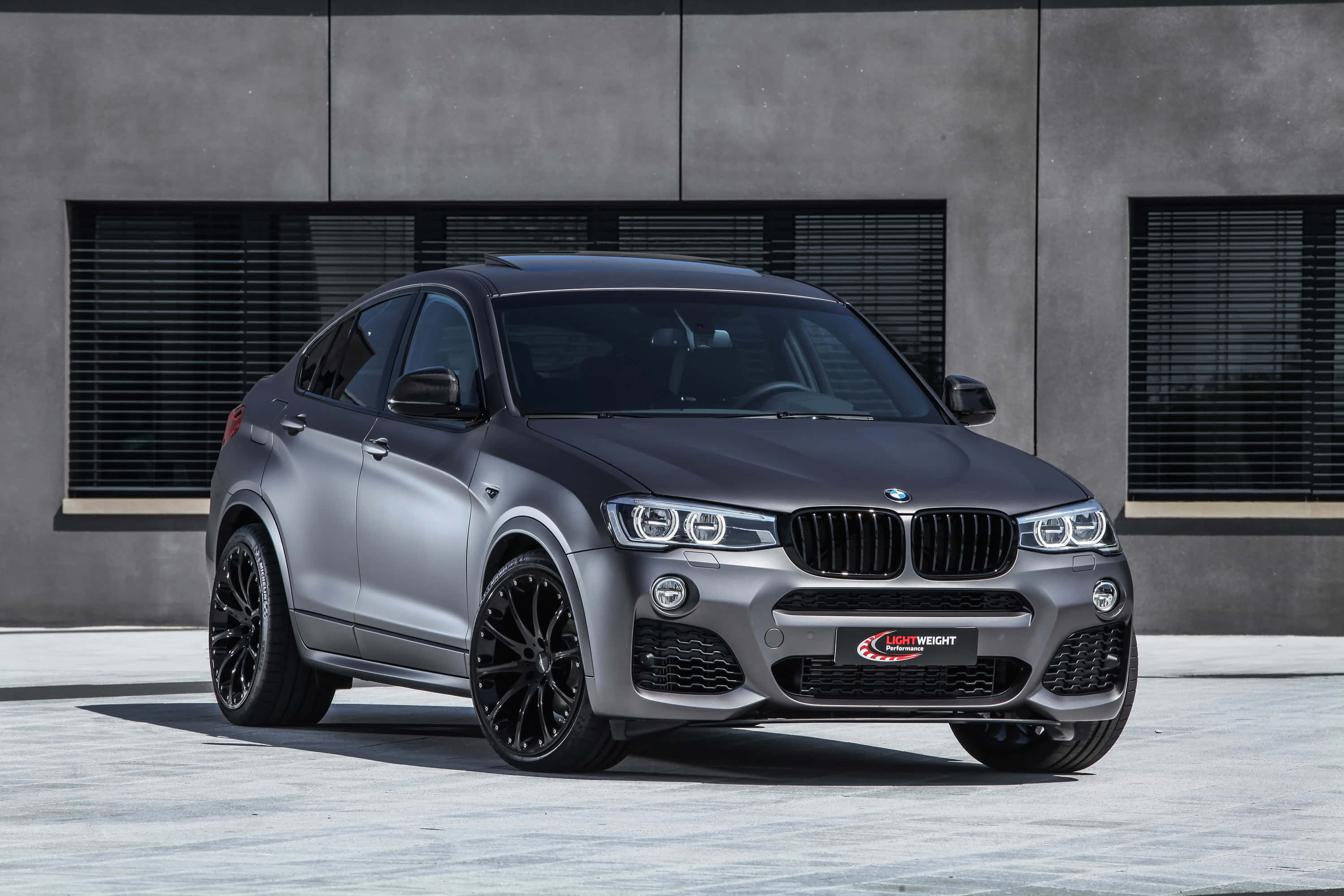 Bmw X4 桌布 壁紙 — 一輛時尚的啞光灰色BMW X4配黑色輪圈，在明亮的日光下停在一棟現代混凝土建築前。