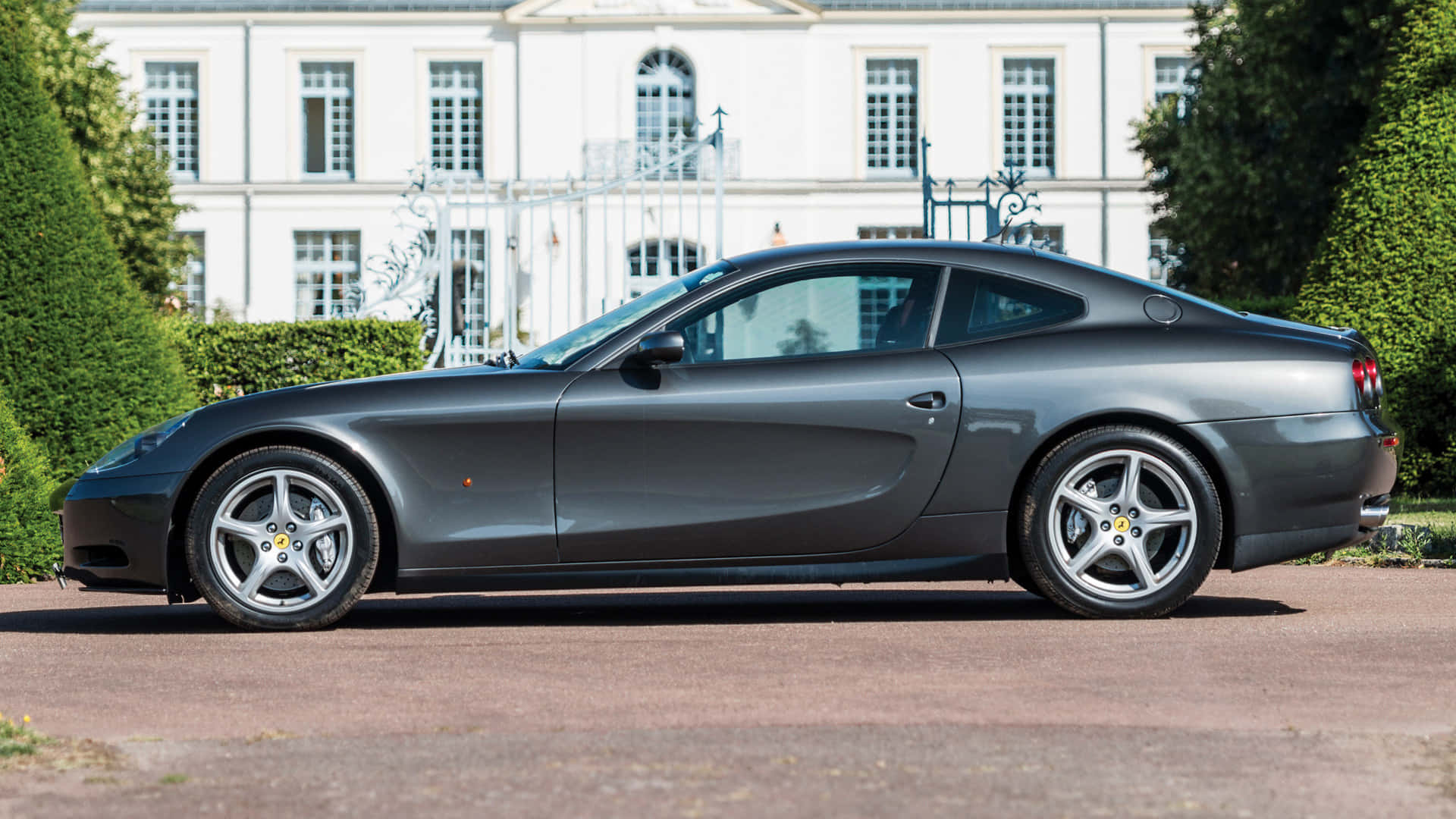 Ferrari 612 Scaglietti 桌布 壁紙 — 一輛時尚的啞光灰色法拉利612 Superamerica停泊在一座帶有修剪整齊樹籬的白色大宅前的碎石車道上。