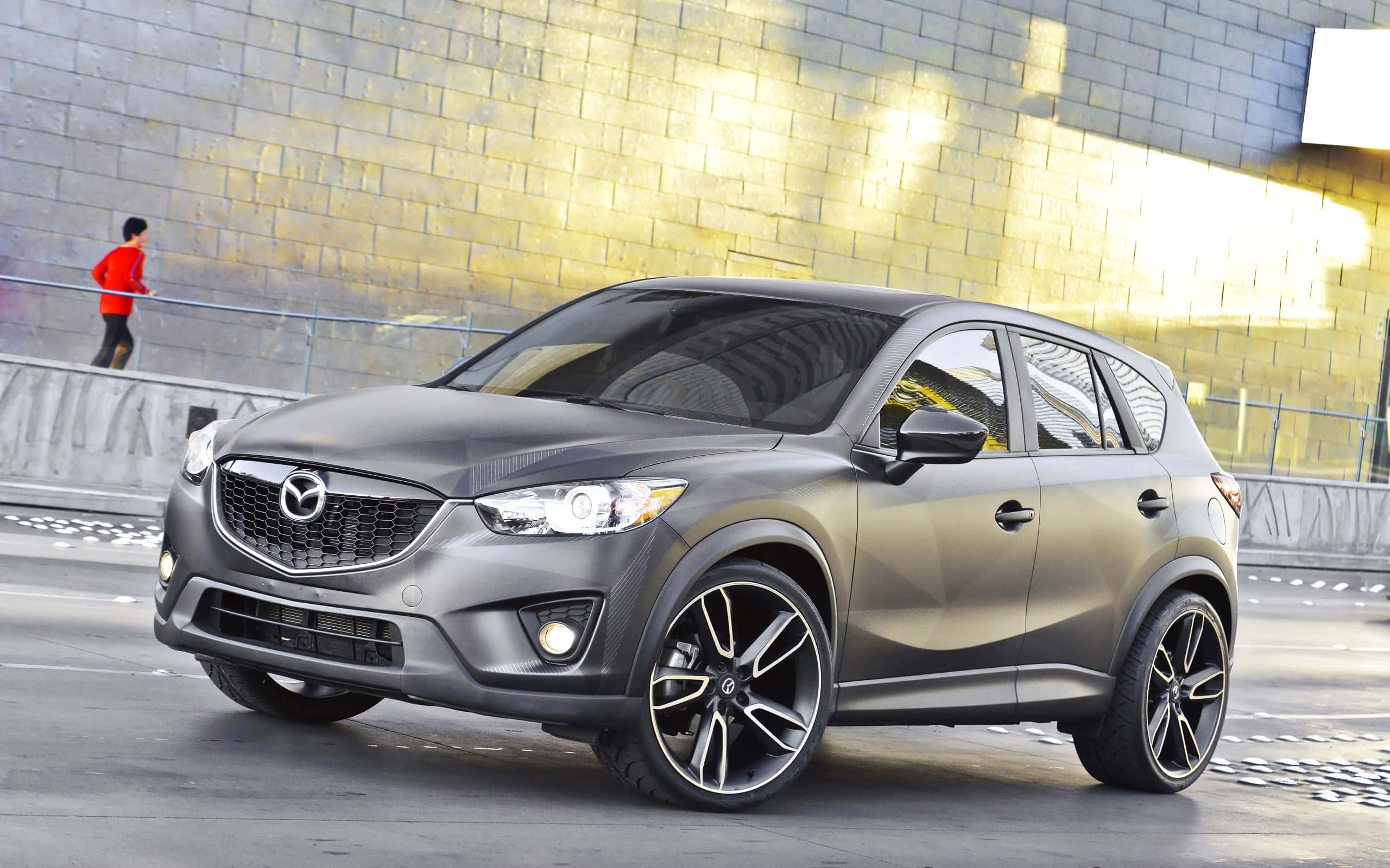 Mazda Cx-5 桌布 壁紙 — 一輛時尚的啞光灰色萬事得CX-5 SUV，配備黑色輪圈，停泊在城市的街道上，旁邊是金色磚牆，一位身穿紅色的跑步者經過。