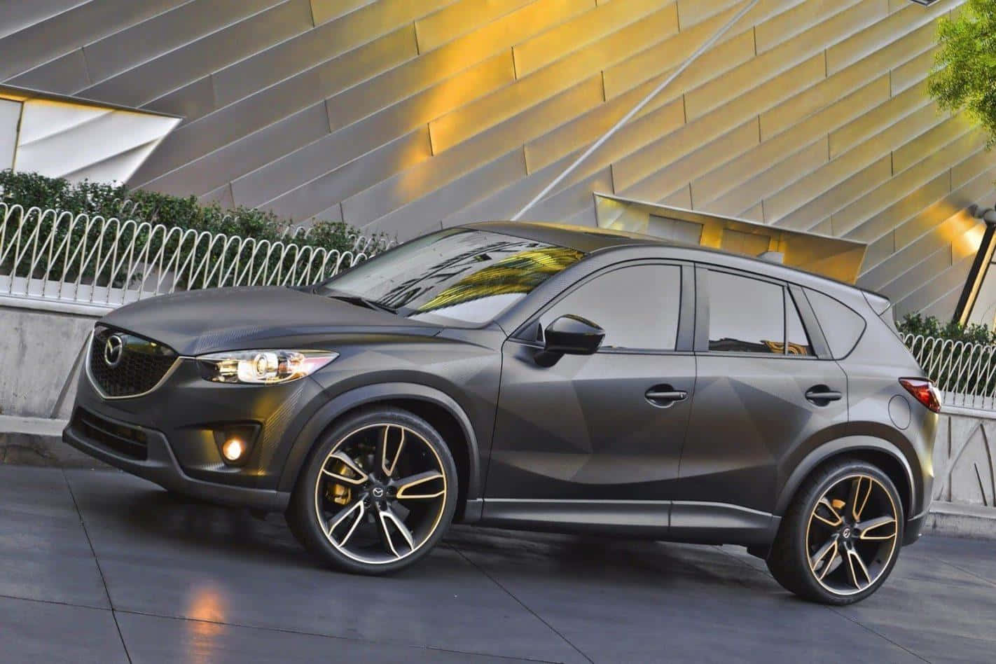 Mazda Cx-5 桌布 壁紙 — 一輛時尚的啞光灰色萬事得CX-5 SUV，配備金色輪圈，停在一個現代棱角建築前，建築帶有溫暖的燈光。