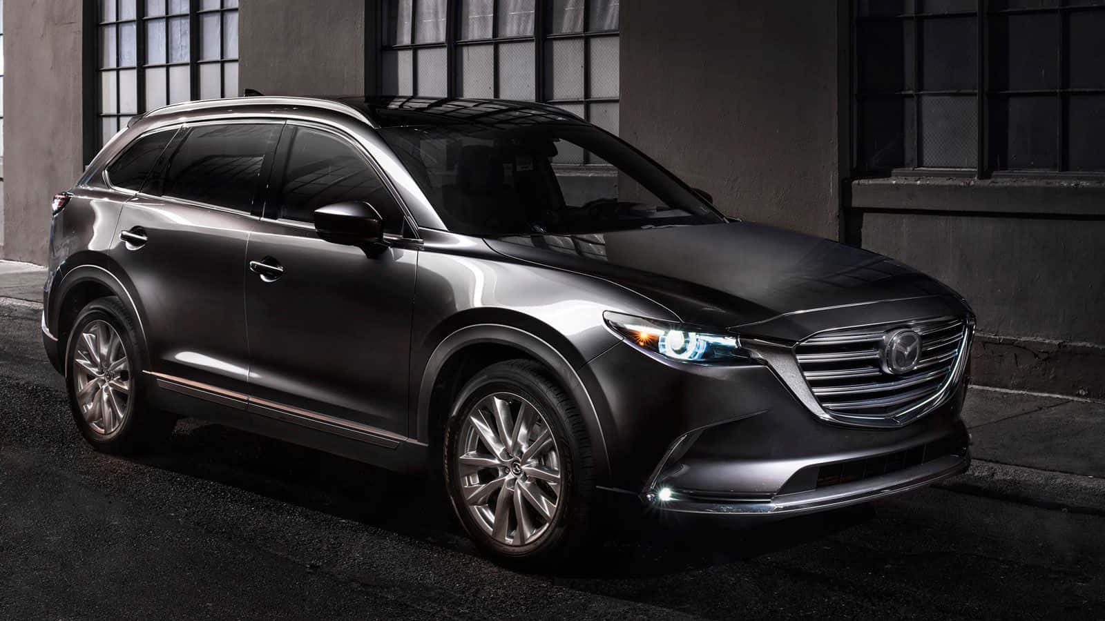 Mazda Cx-9 桌布 壁紙 — 一輛時尚的啞光灰色萬事達CX-9 SUV在夜間停在一棟有大窗戶的建築物前。