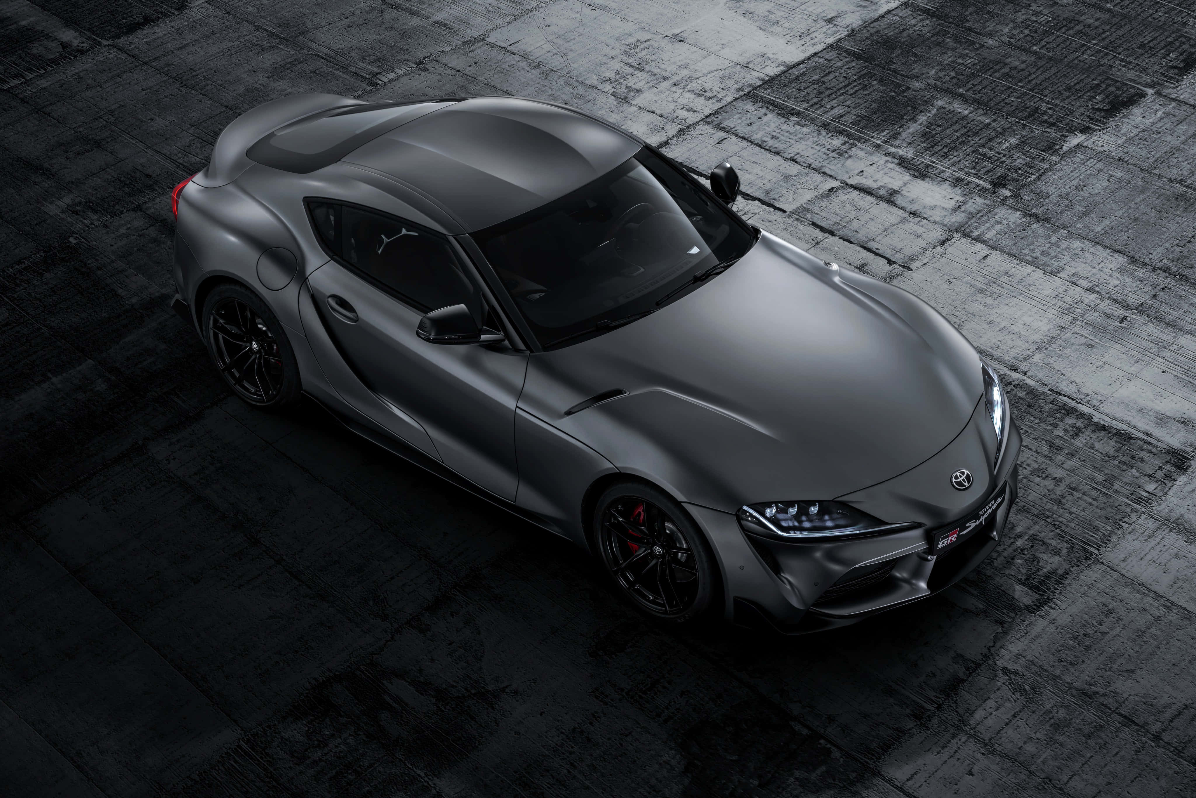Supra 桌布 壁紙 — 一輛時尚的啞光灰色跑車，帶有黑色飾件和紅色制動卡鉗，在戲劇性燈光下停放在深色紋理混凝土表面上。