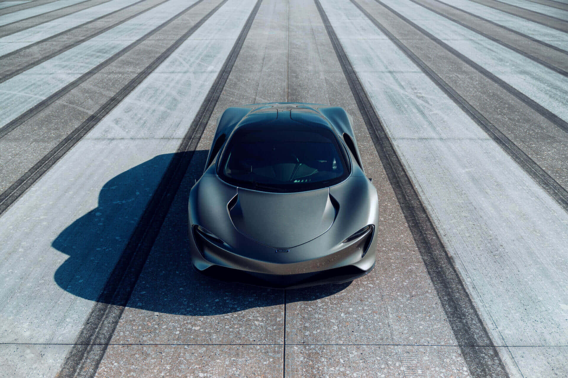 Mclaren Speedtail 桌布 壁紙 — 一輛時尚的啞光灰色跑車在明亮的日光下，居中停放在一條帶有平行輪胎痕的混凝土賽道上。