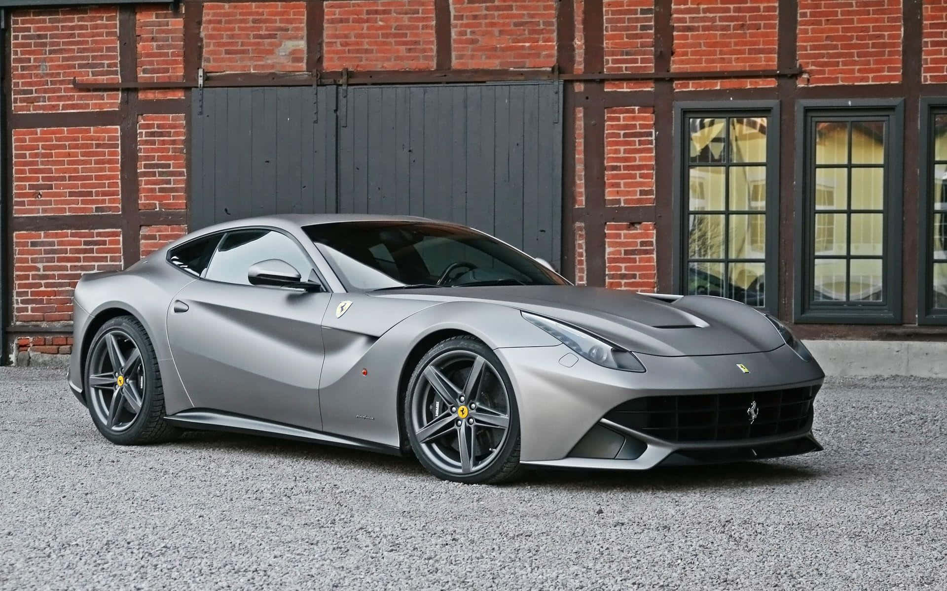 Ferrari F12 Berlinetta 桌布 壁紙 — 一輛時尚的啞光銀色法拉利F12 Berlinetta停在碎石路上，前面是一座帶有深色木框架的質樸紅磚建築。