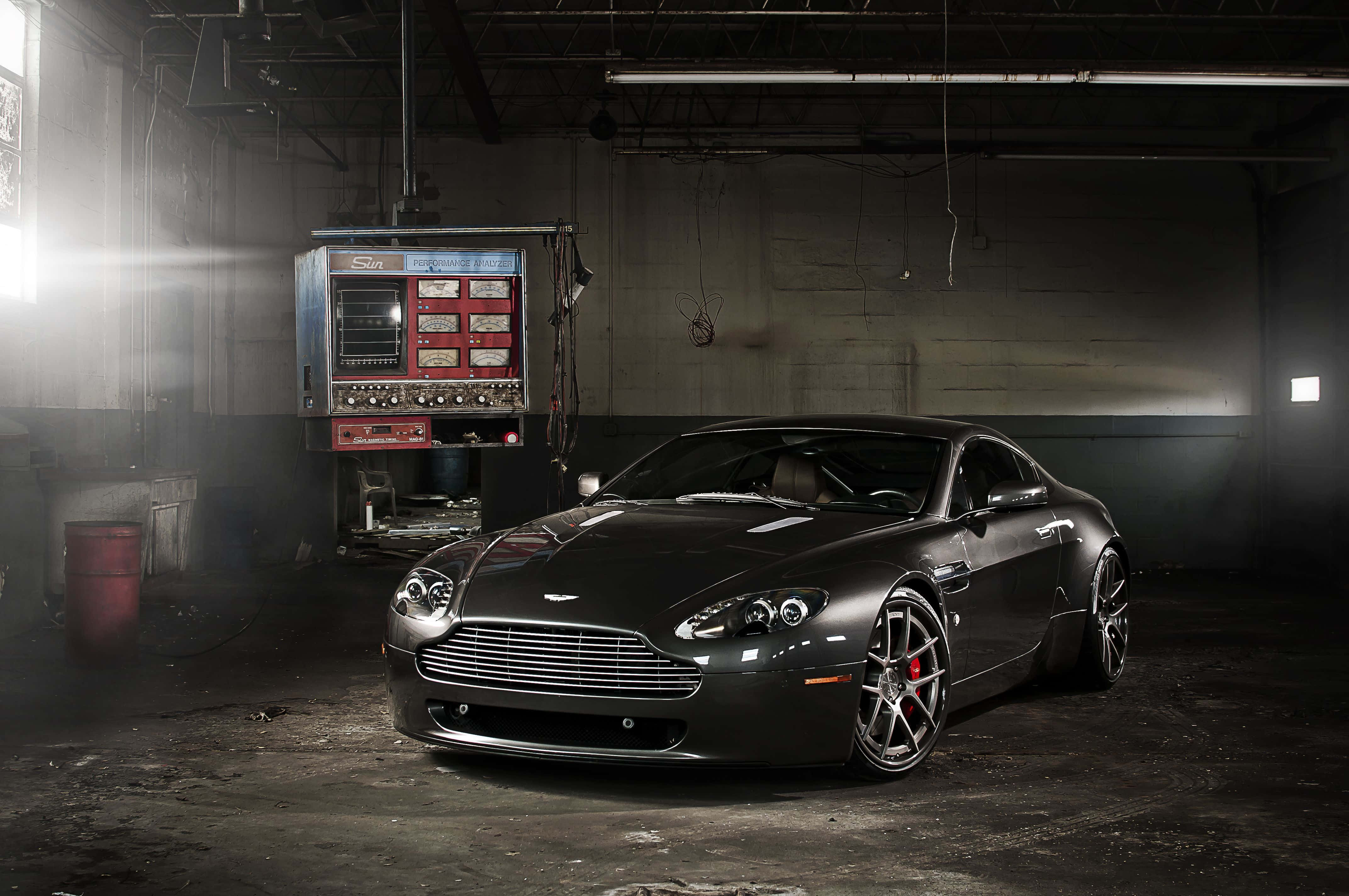 Aston Martin V12 Vantage 桌布 壁紙 — 一輛時尚的啞光黑色Aston Martin跑車在粗獷的工業車庫中，在戲劇性的聚光燈下閃閃發光。