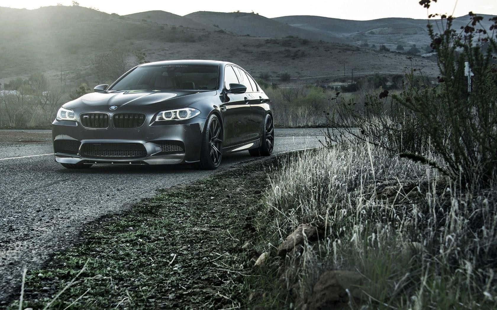 Bmw M5 桌布 壁紙 — 一輛時尚的啞光黑色BMW M5運動轎車停在碎石路上，背景是黃昏時分起伏的山丘和乾草。