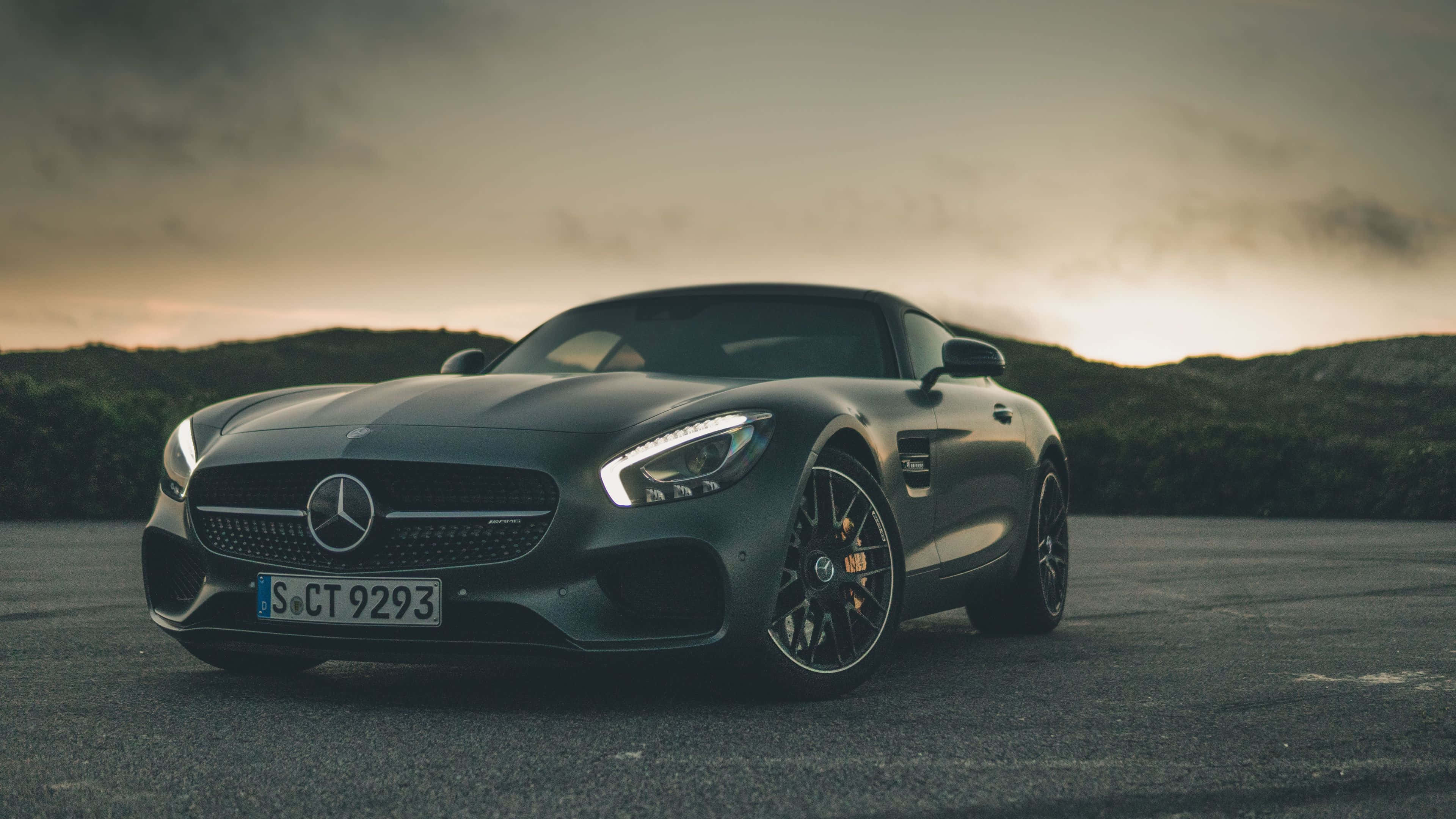4K Mercedes 桌布 壁紙 — 一輛時尚的啞光黑色Mercedes-AMG GT跑車停在瀝青路上，背後是起伏山丘上戲劇性的日落天空。