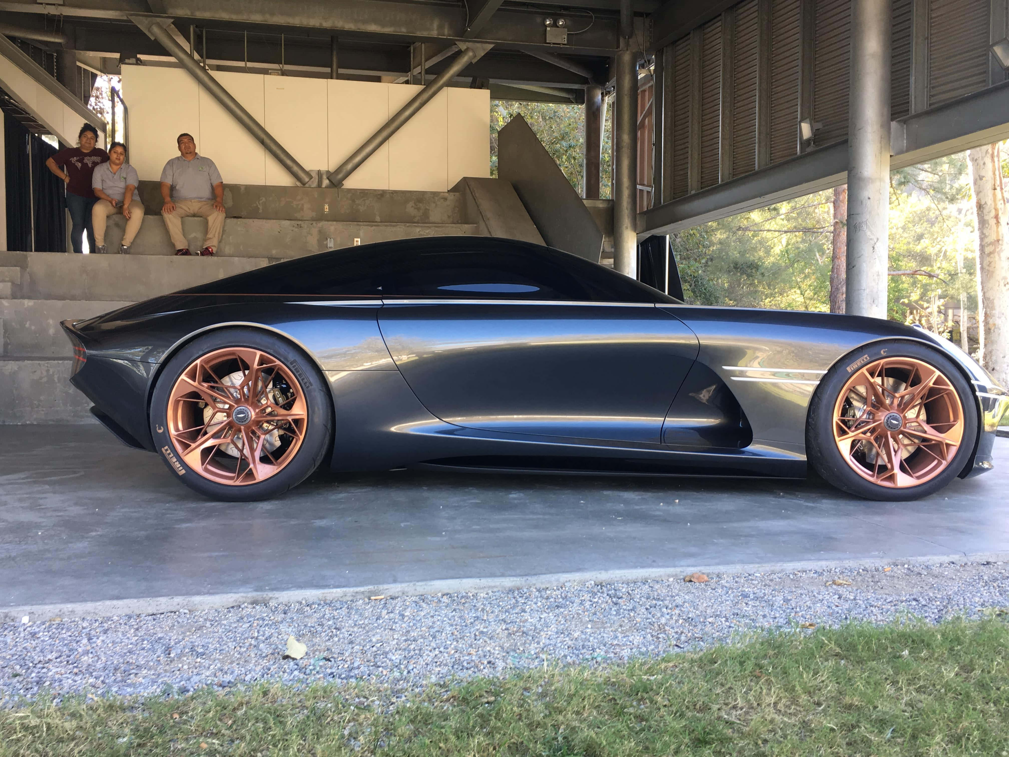 Genesis Essentia 桌布 壁紙 — 一輛時尚的啞光黑色概念車，配備銅色車輪，停放在現代混凝土結構下，有三名男子在觀察。