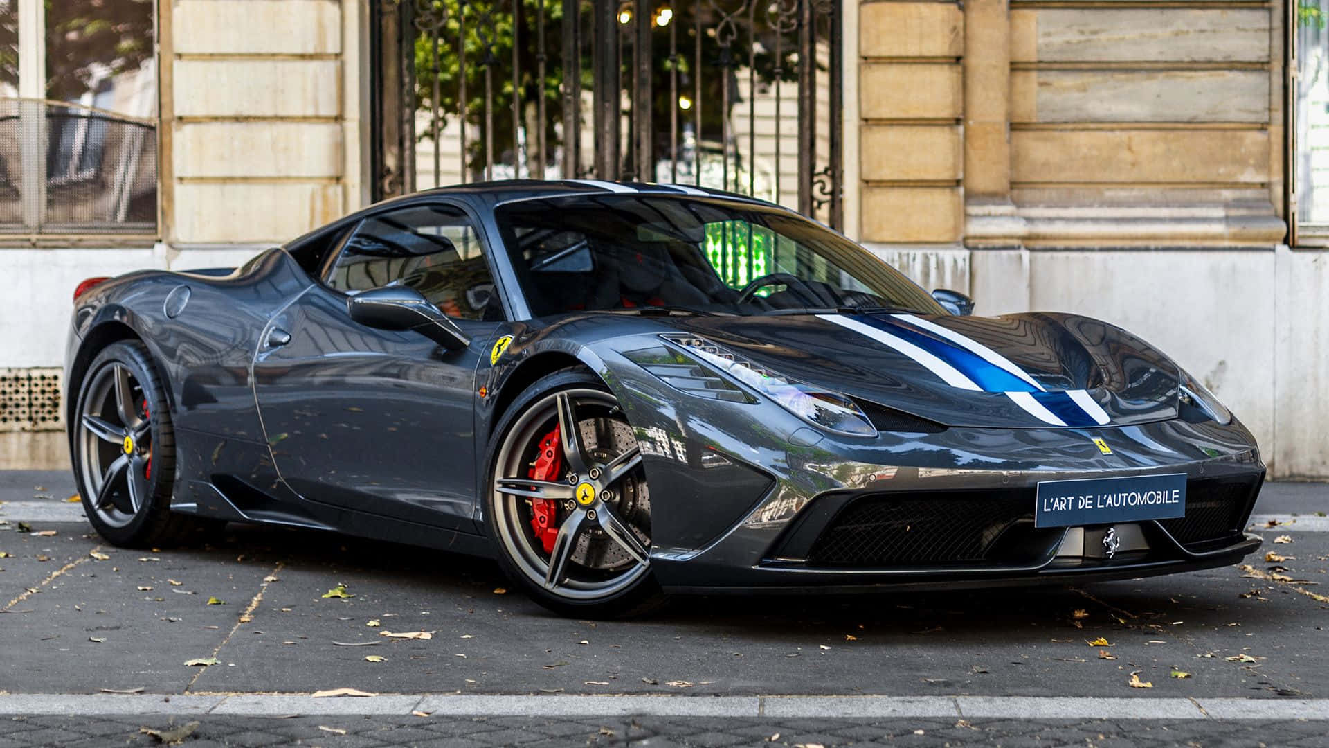 Ferrari 458 Speciale 桌布 壁紙 — 一輛時尚的啞光黑色法拉利跑車，帶有藍色賽車條紋，停在歷史建築旁邊的城市街道上。