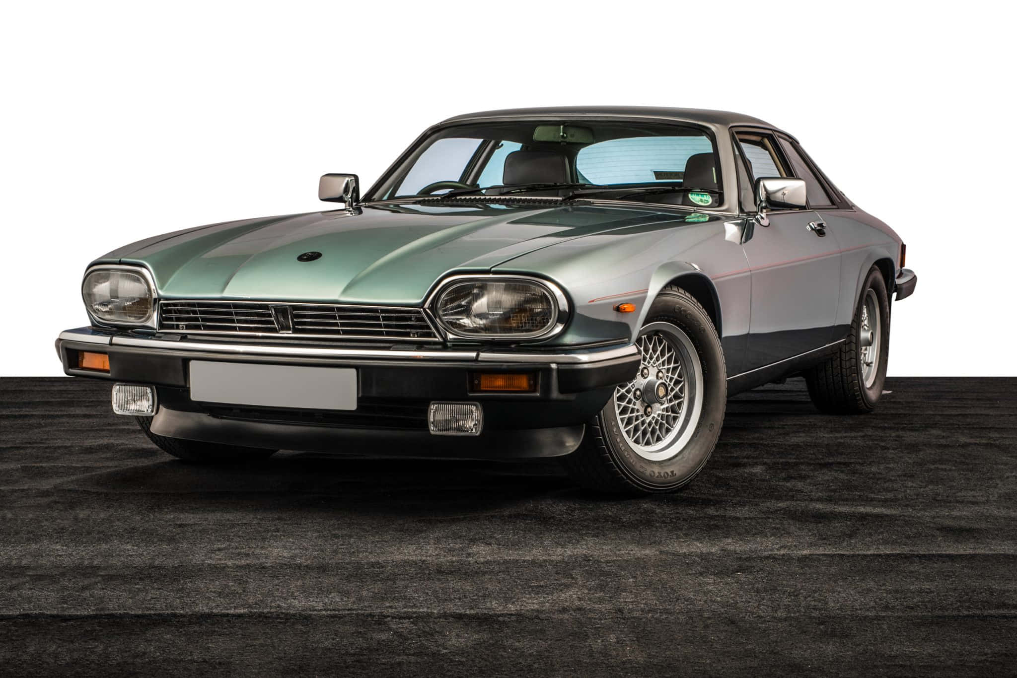 Jaguar Xjs 桌布 壁紙 — 一輛時尚的復古捷豹XJ-S跑車，金屬綠色，帶有鍍鉻裝飾和鋼絲輪，放置在白色背景前的深色紋理地板上。