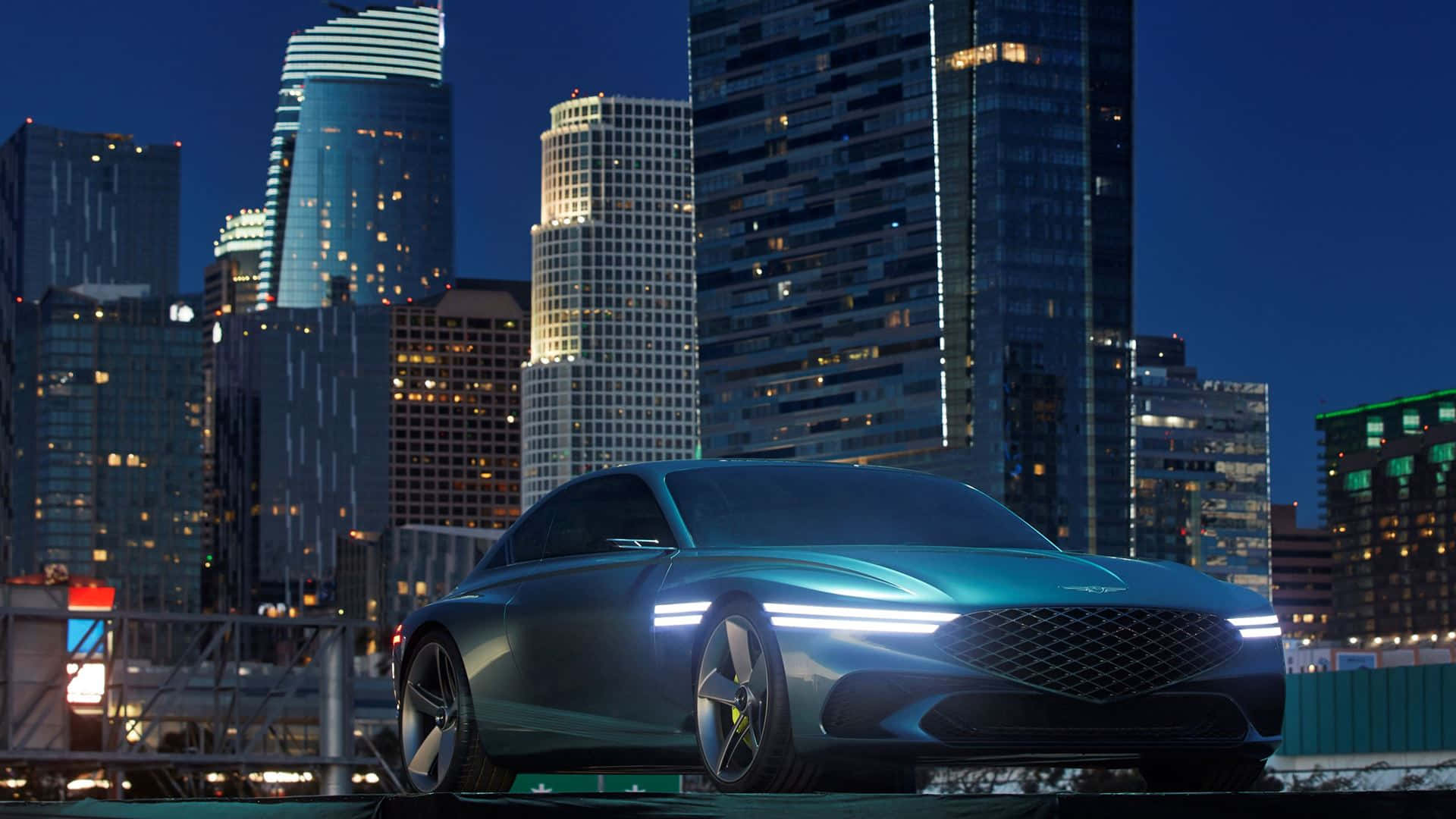 Genesis X Concept 桌布 壁紙 — 一輛時尚的未來感青綠色汽車在暮色中，以亮起的車頭燈映襯著高聳的城市天際線。