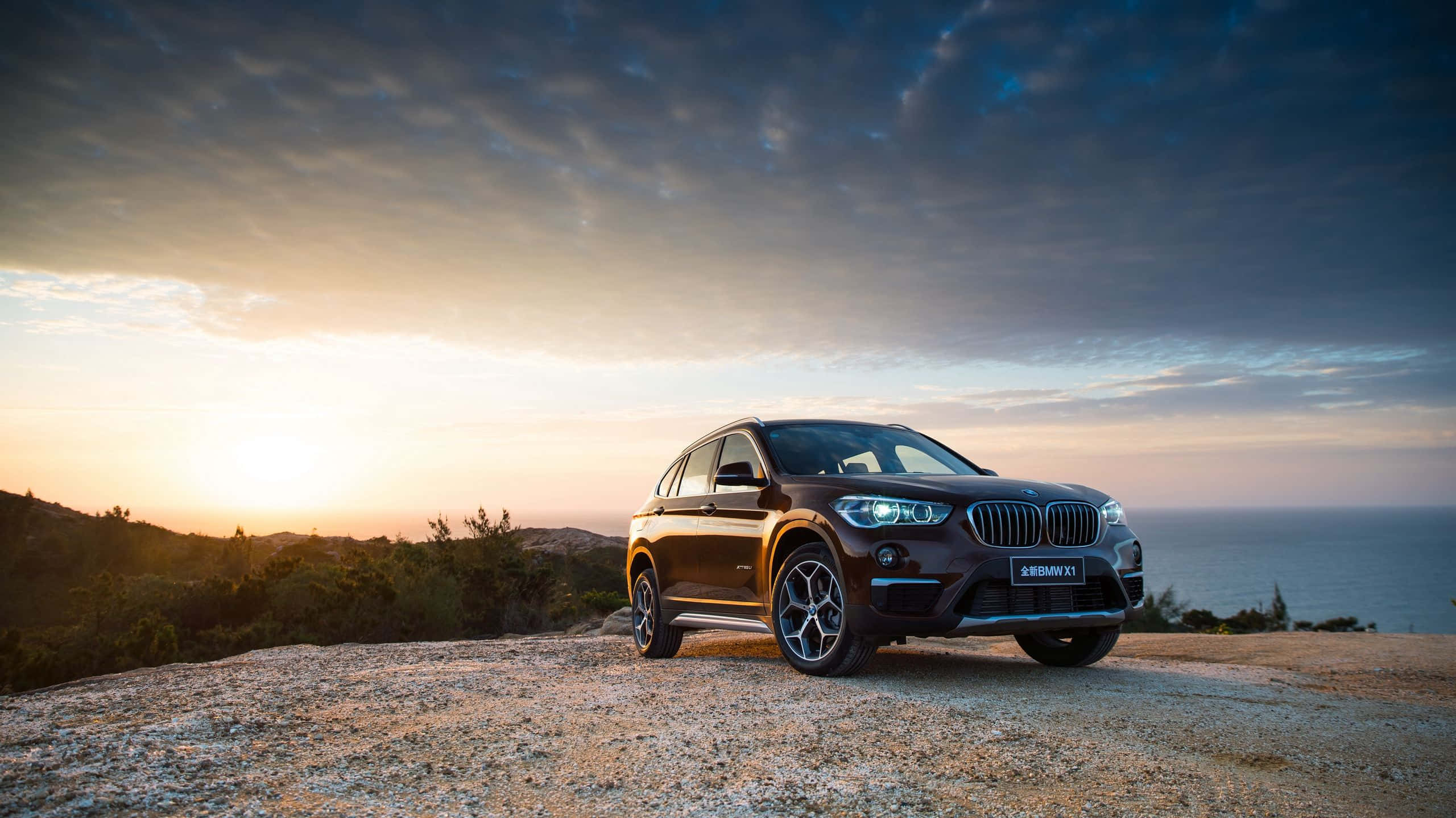 Bmw X1 桌布 壁紙 — 一輛時尚的棕色BMW X1 SUV停泊在岩石懸崖上，俯瞰日落時分的海洋，天空中有戲劇性的雲彩。