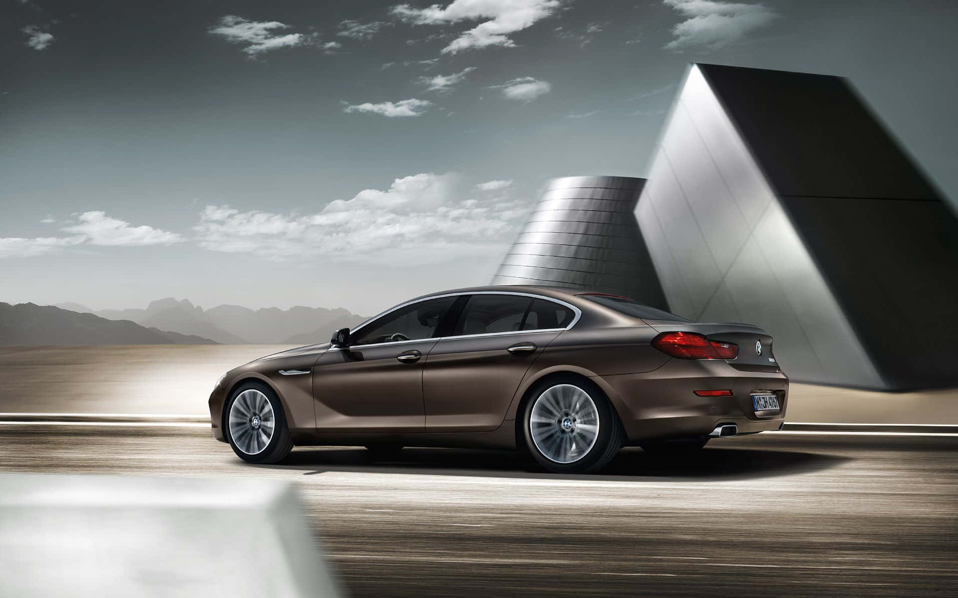 Bmw 6 Series 桌布 壁紙 — 一輛時尚的棕色BMW房車在陰天和遠山的襯托下滑過一座未來主義的金屬建築。
