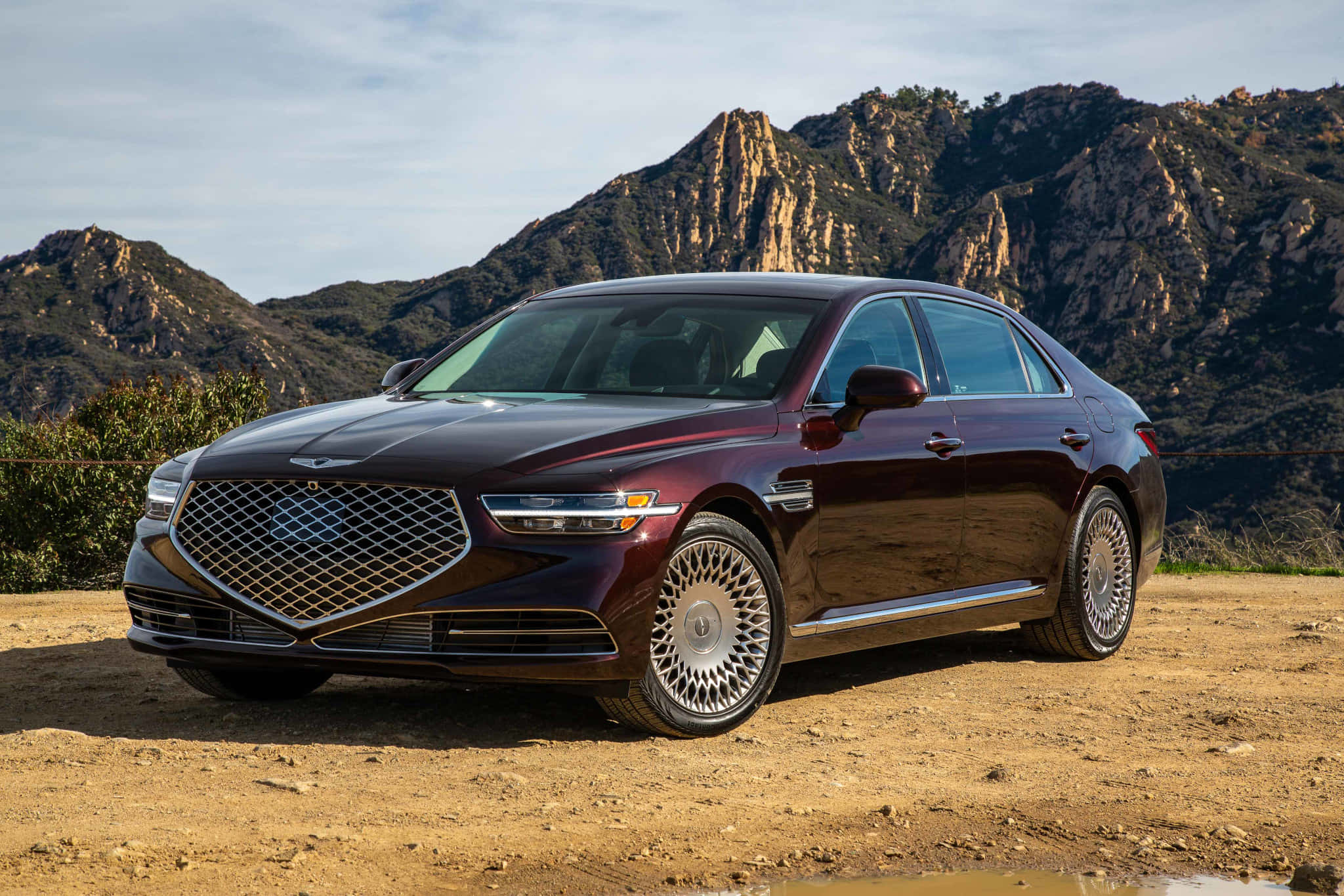 Genesis G90 桌布 壁紙 — 一輛時尚的棗紅色 Genesis G90 轎車停泊在塵土飛揚的地形上，背後是崎嶇的山脈，在晴朗的天空下。