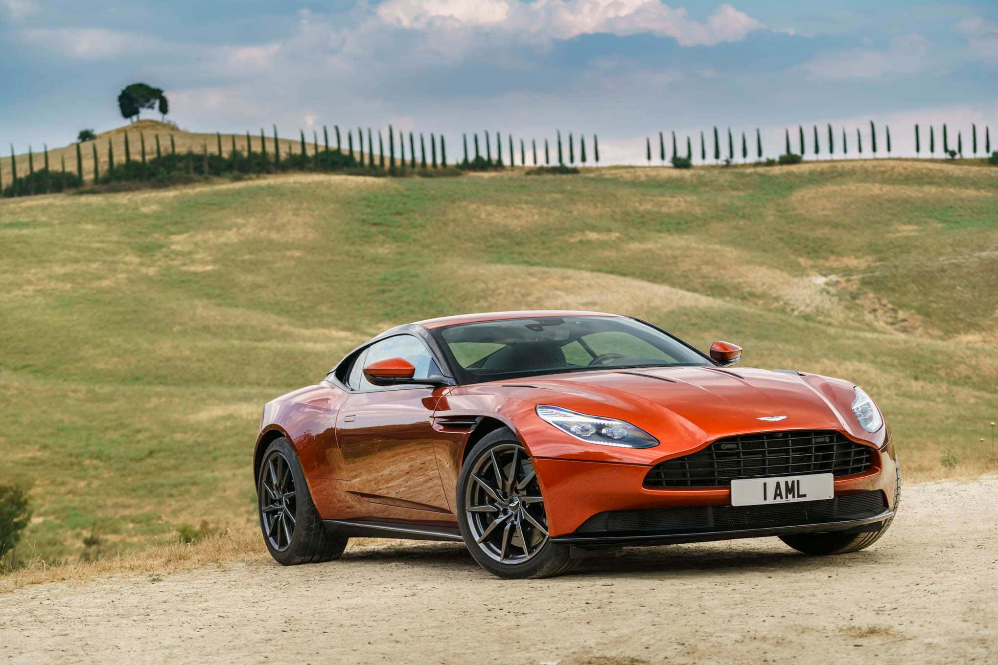 Aston Martin Db11 桌布 壁紙 — 一輛時尚的橙色Aston Martin跑車停泊在土路上，周圍是起伏的托斯卡納山丘，柏樹林立，天空部分多雲。