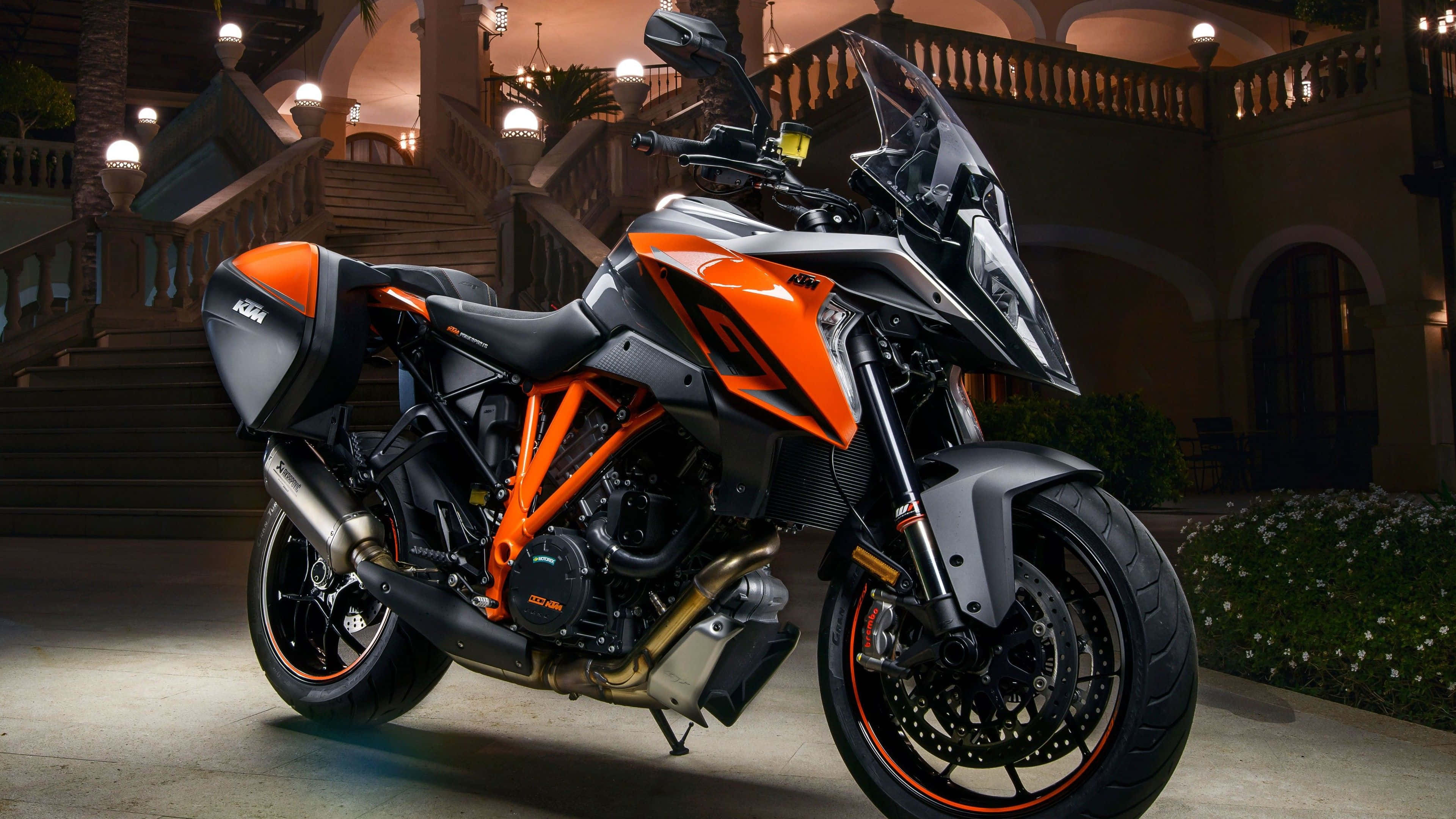Ktm Bike 桌布 壁紙 — 一輛時尚的橙色和黑色KTM電單車在夜間停泊在一座宏偉的照明建築前，該建築有石階和陽台。