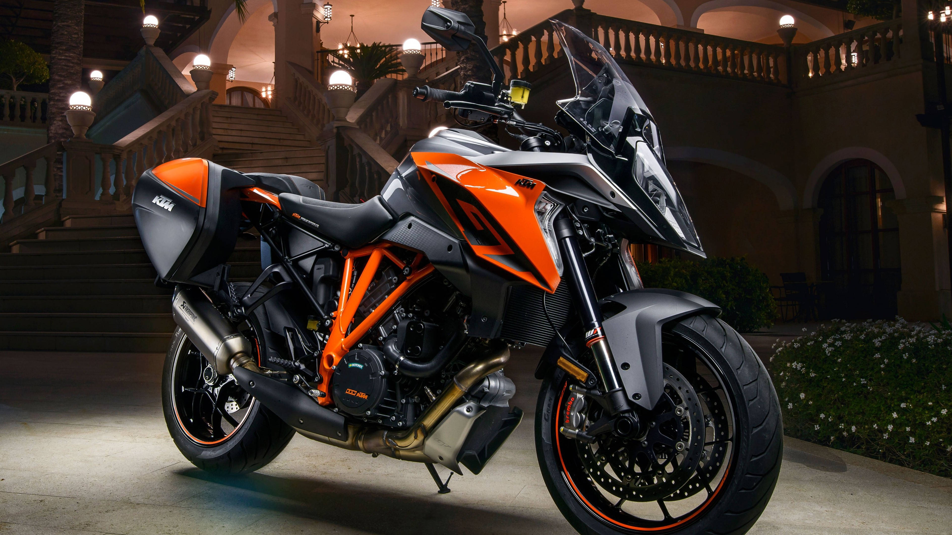 ktm bikes 桌布 壁紙 — 一輛時尚的橙色和黑色KTM電單車在夜間停放在宏偉的石階上，周圍有溫暖的環境照明。