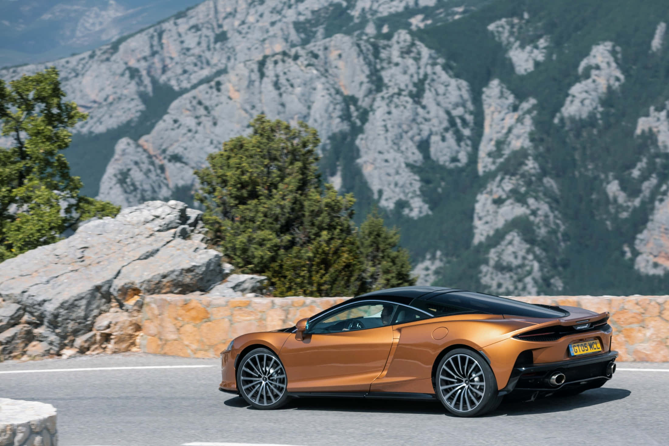 Mclaren Gt 桌布 壁紙 — 一輛時尚的橙色跑車沿著山路行駛，背景是崎嶇的山峰和綠樹。