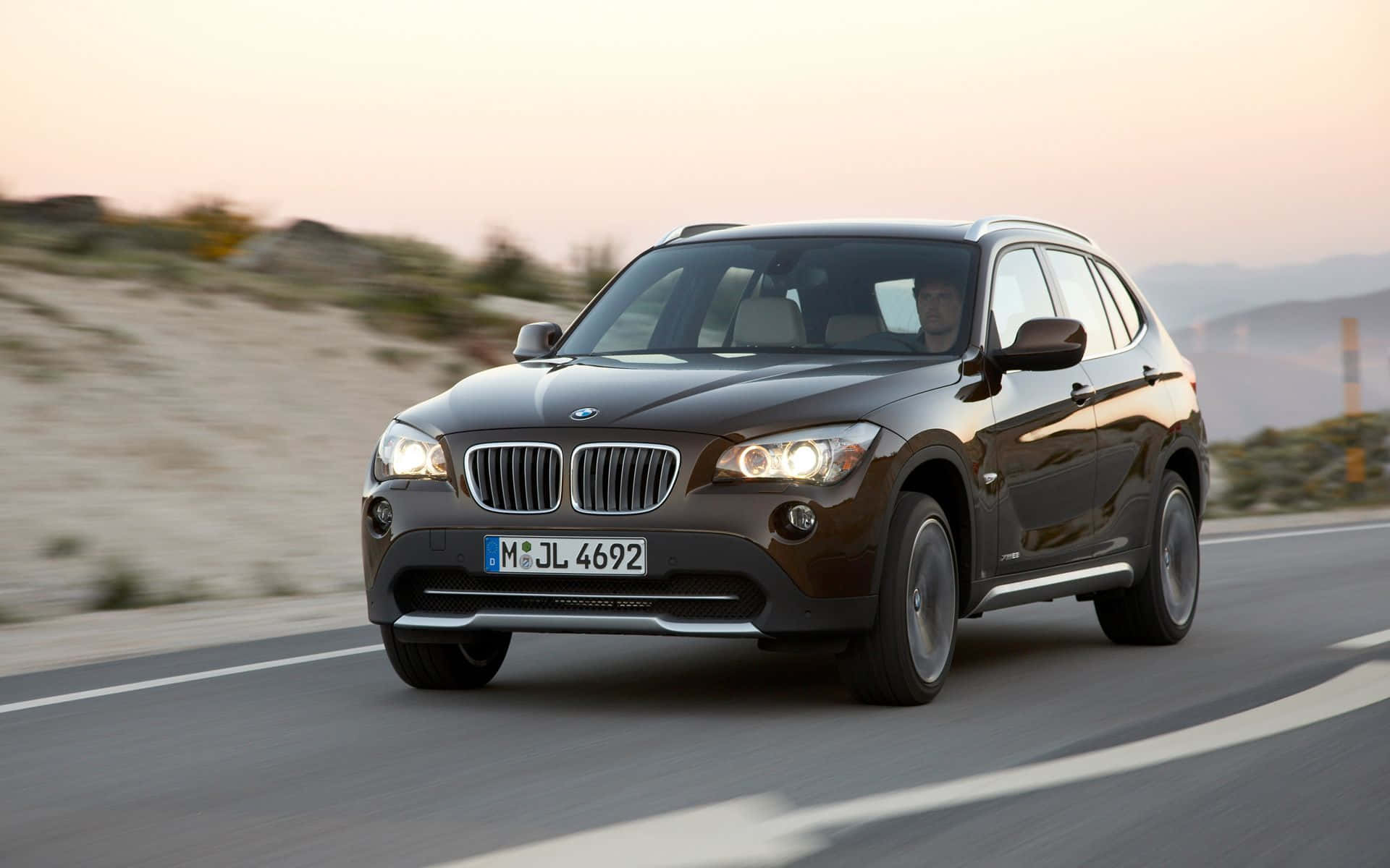 Bmw X1 桌布 壁紙 — 一輛時尚的深棕色BMW X1 SUV在日落時分沿著蜿蜒的海岸公路行駛，帶有模糊的動態和溫暖的金色光線。