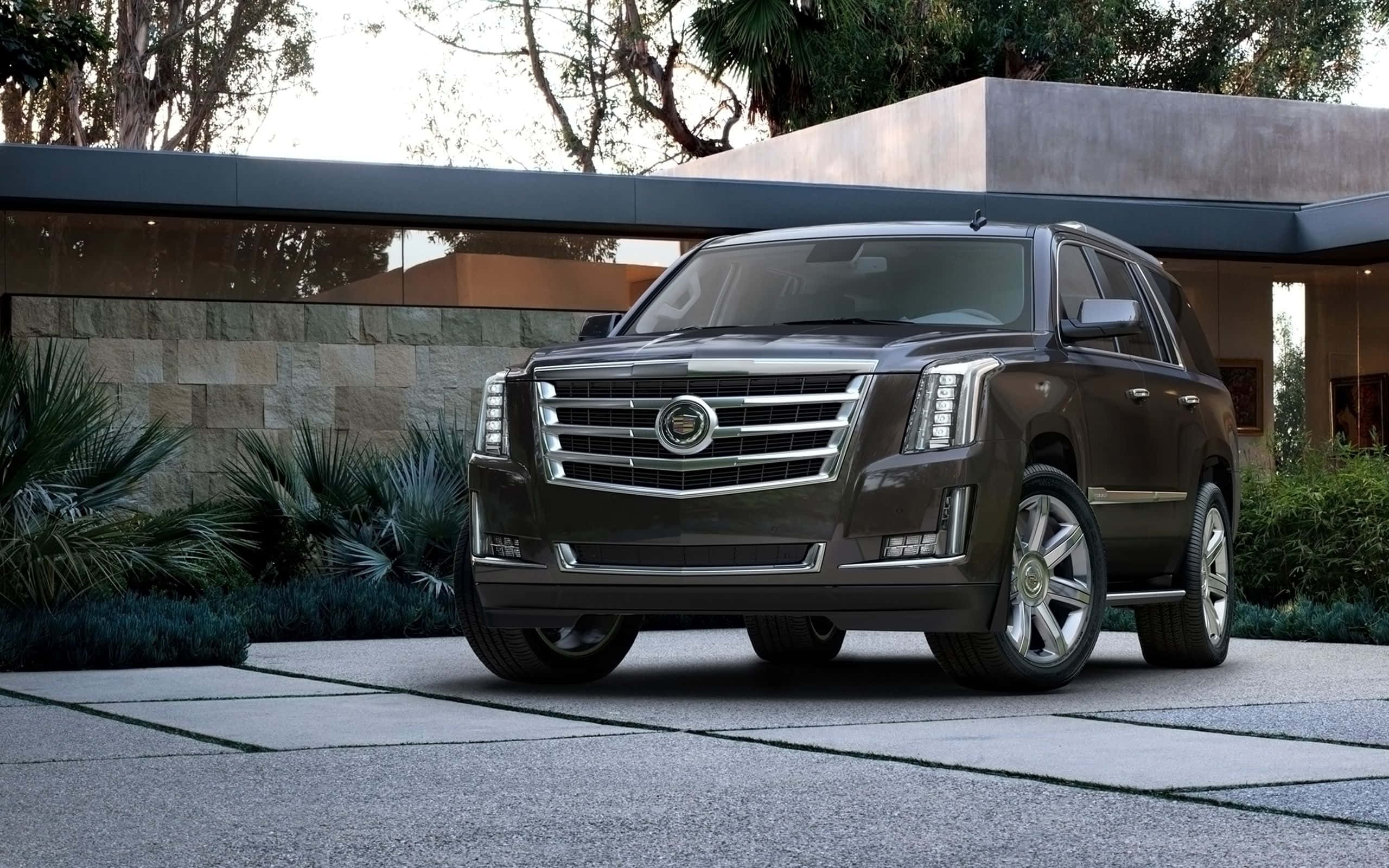 Cadillac Escalade 桌布 壁紙 — 一輛時尚的深棕色Cadillac SUV停泊在現代石頭車道上，前方是一座擁有茂密綠植的當代房屋。