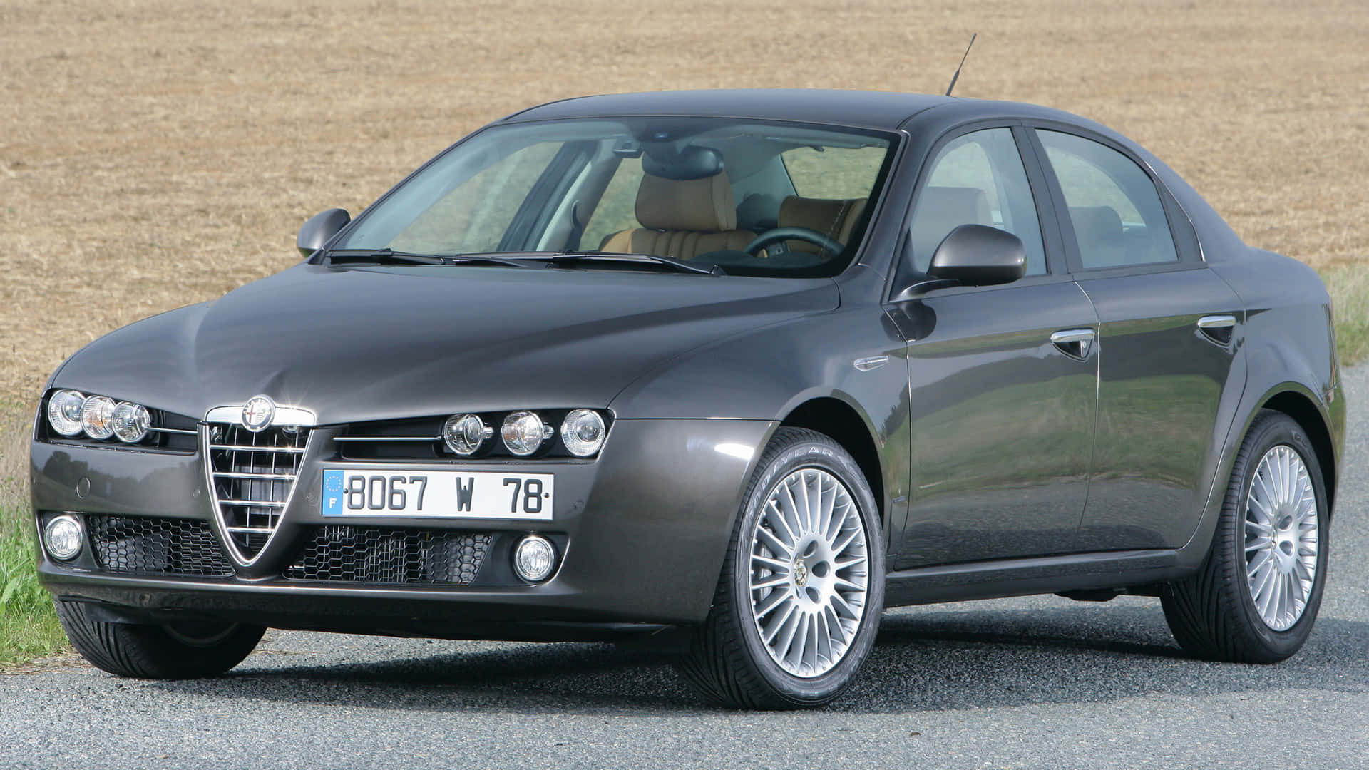 Alfa Romeo 159 桌布 壁紙 — 一輛時尚的深灰色 Alfa Romeo 房車停在碎石上，後面是乾燥的田野。