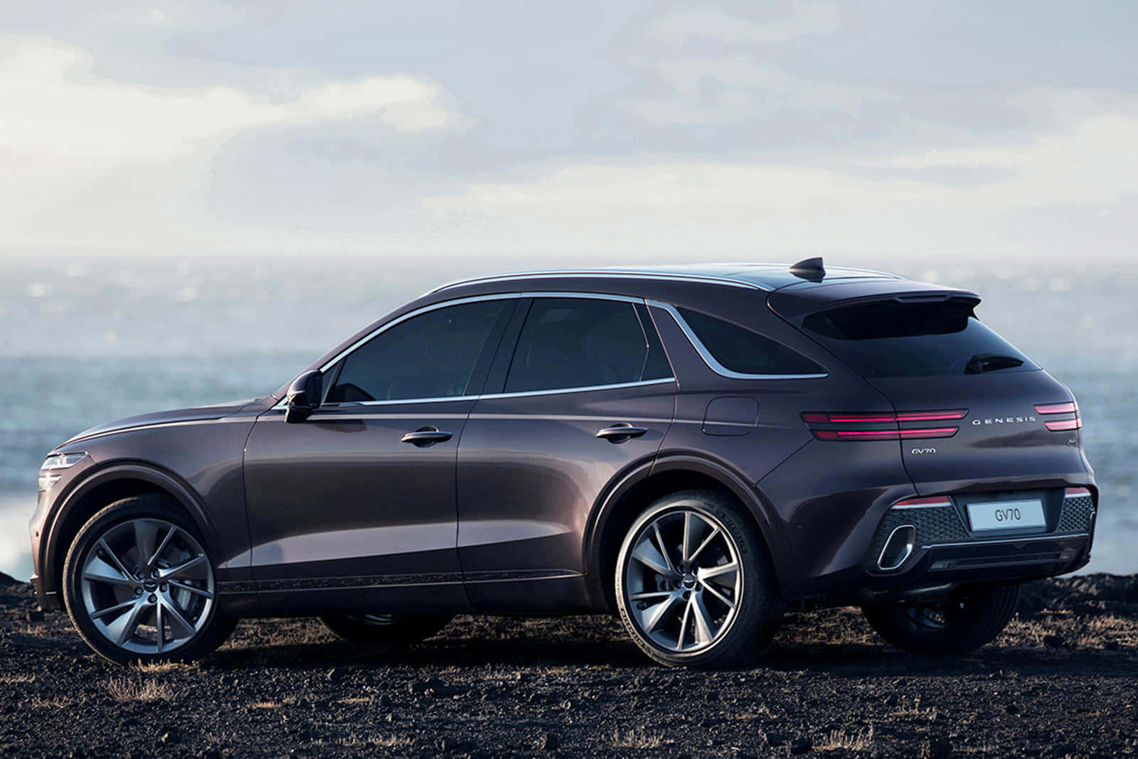 Genesis Gv70 桌布 壁紙 — 一輛時尚的深灰色 Genesis GV70 SUV 停泊在黑色火山沙上，旁邊是平靜的海洋，天空柔和多雲。