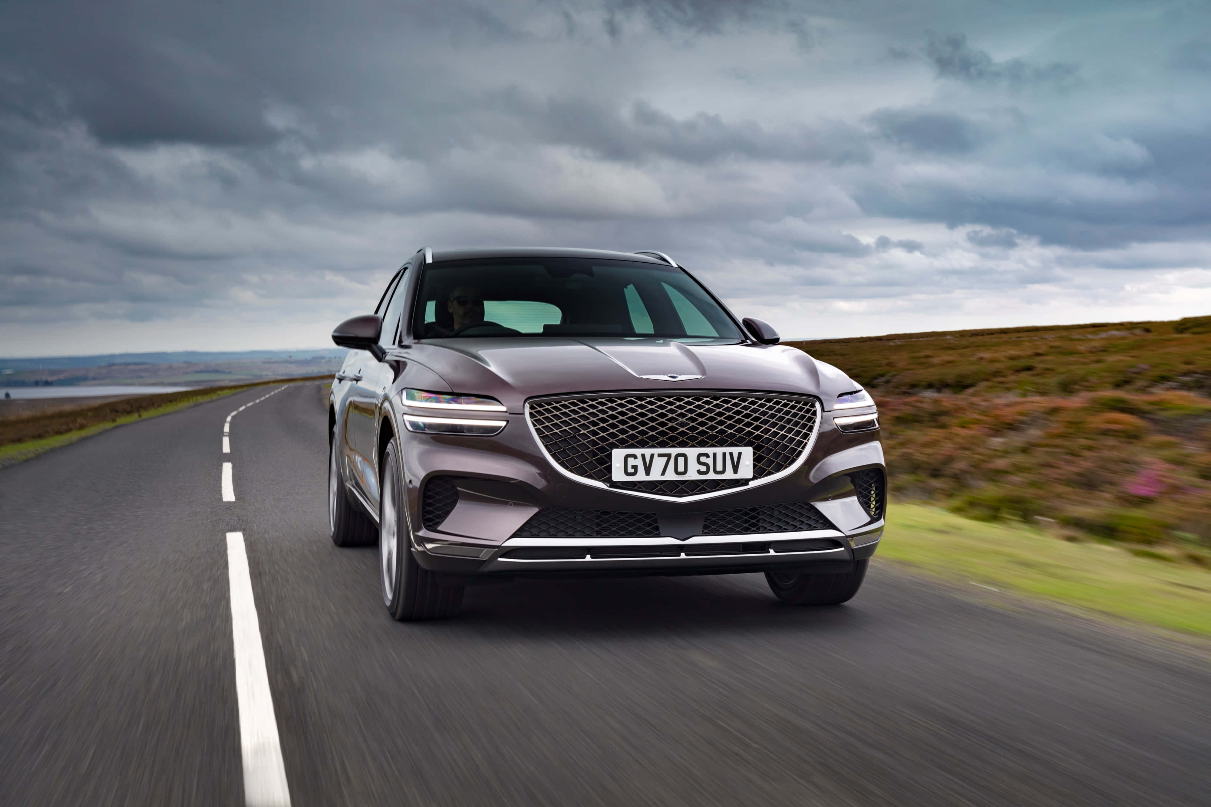 Genesis Gv70 桌布 壁紙 — 一輛時尚的深灰色 Genesis GV70 SUV 在戲劇性的多雲天空下，沿著蜿蜒的海岸道路飛馳。