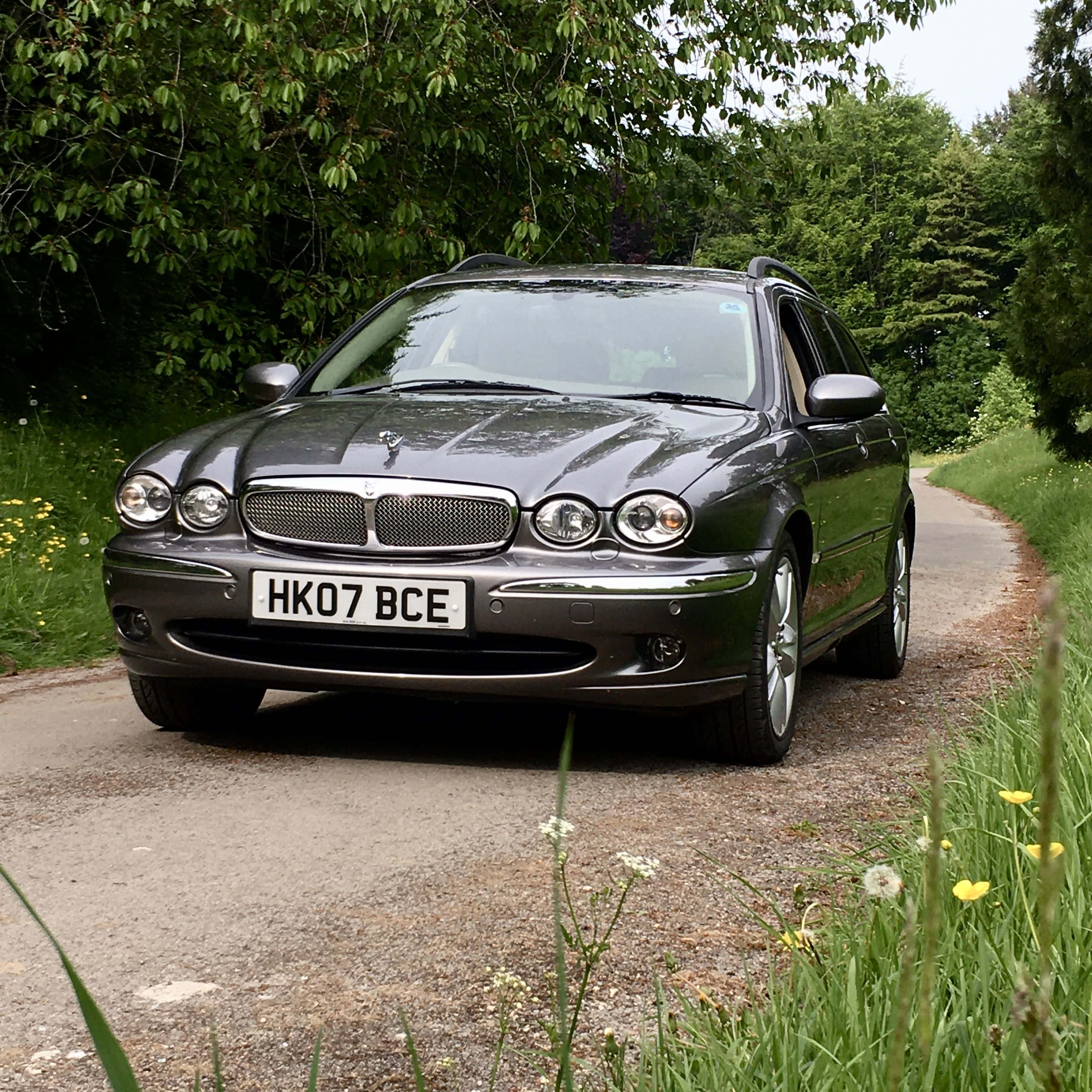 Jaguar X-type 桌布 壁紙 — 一輛時尚的深灰色 Jaguar 旅行車停泊在碎石鄉村道路上，兩旁是茂密的綠樹和野花。