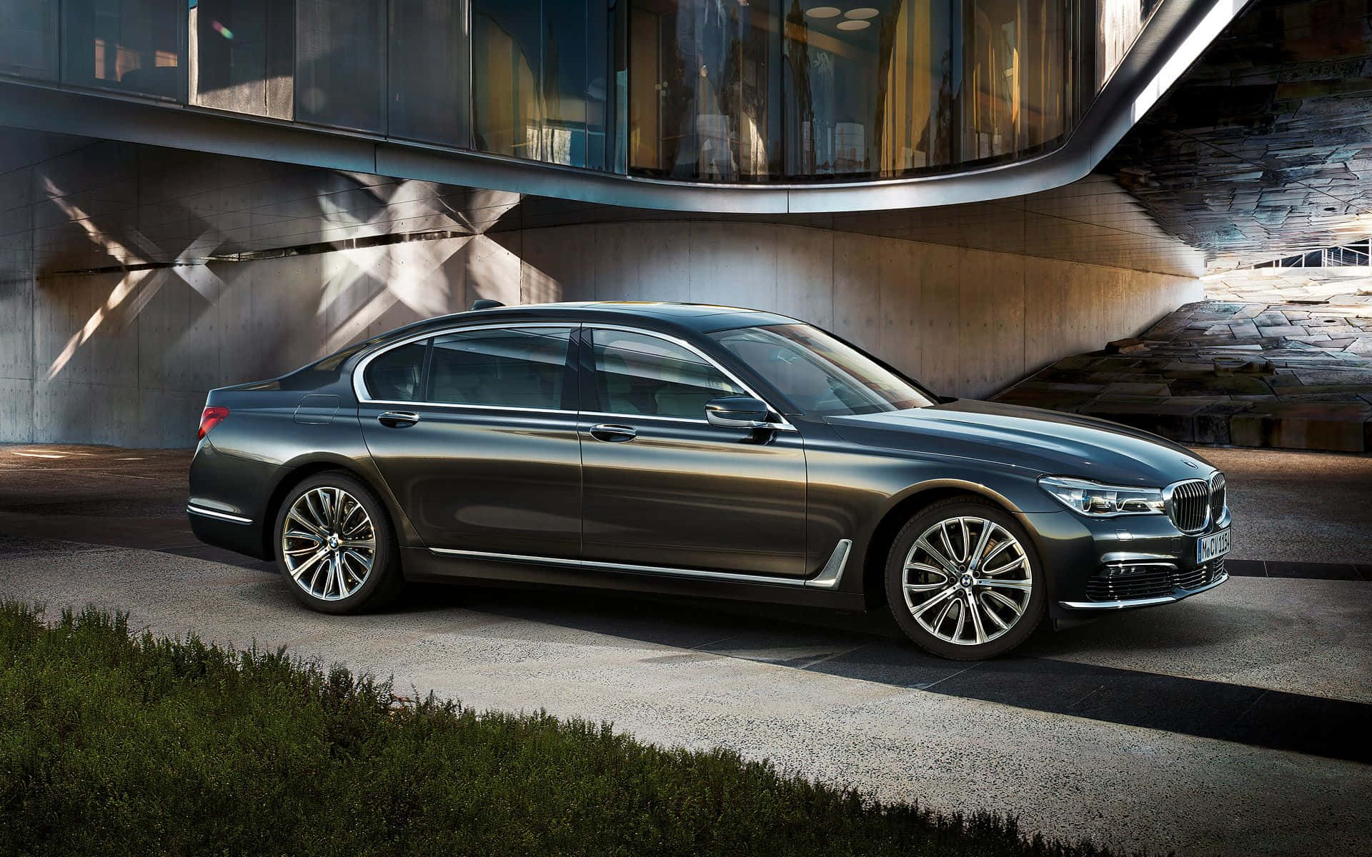 Bmw 7 Series 桌布 壁紙 — 一輛時尚的深灰色BMW 7系轎車停放在現代弧形玻璃和混凝土建築下，戲劇性的陽光投射出幾何陰影。