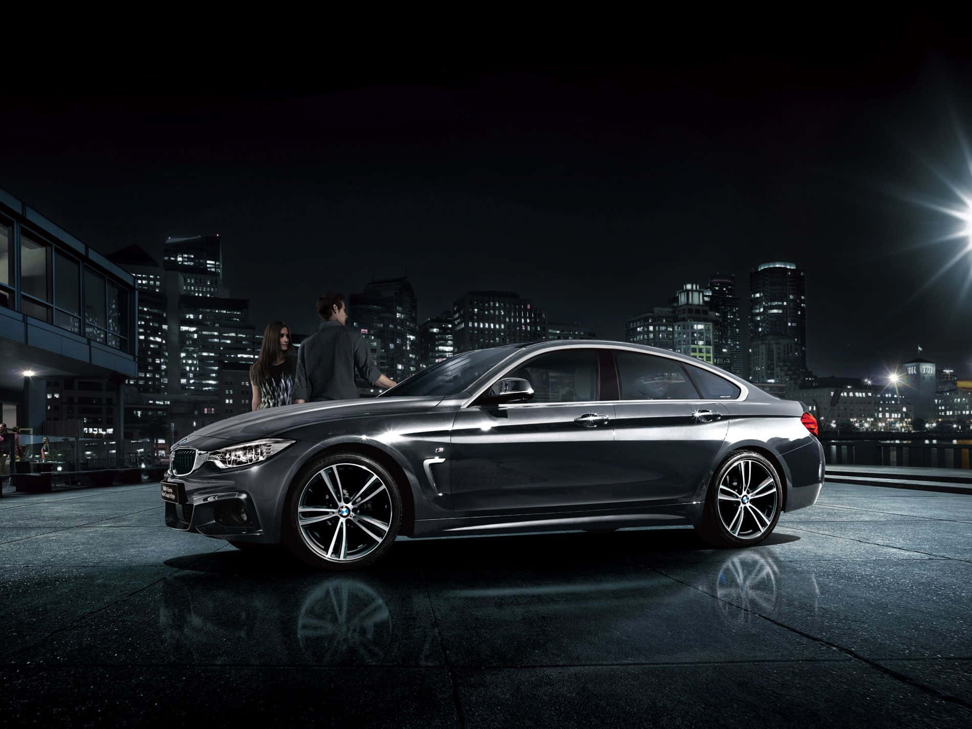 Bmw 4 Series 桌布 壁紙 — 一輛時尚的深灰色BMW房車在城市的燈光下閃閃發光，有兩個人在潮濕的屋頂上欣賞它。