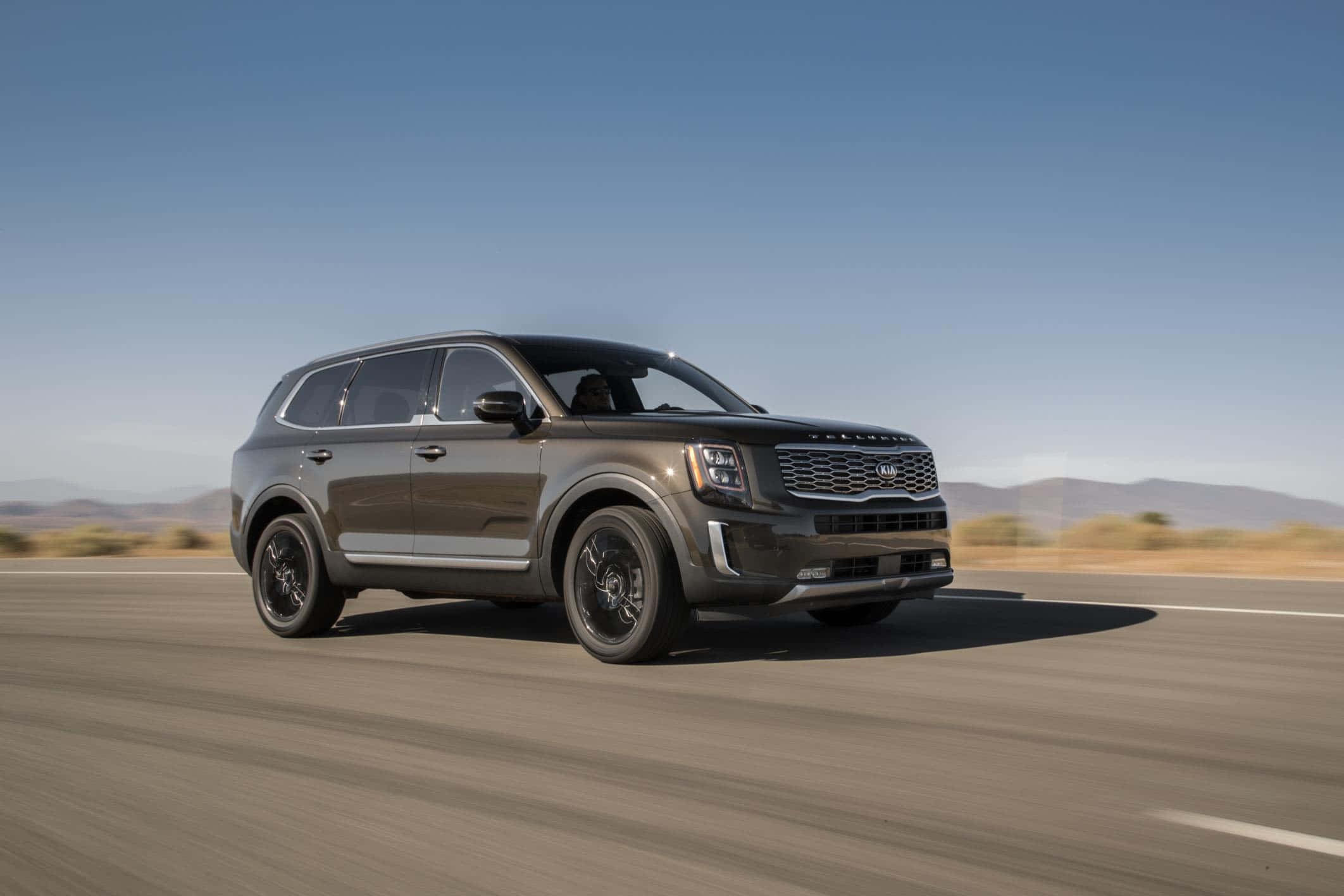 Kia Telluride 桌布 壁紙 — 一輛時尚的深灰色Kia Telluride SUV在晴朗藍天下沿沙漠公路行駛，遠山為背景。