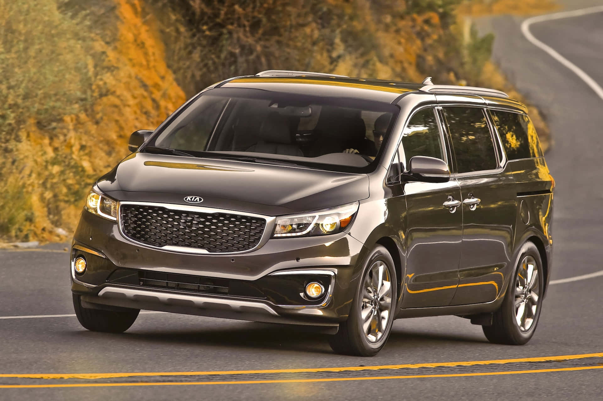 Kia Sedona 桌布 壁紙 — 一輛時尚的深灰色Kia小型貨車沿著蜿蜒的山路行駛，周圍是秋天的樹葉。
