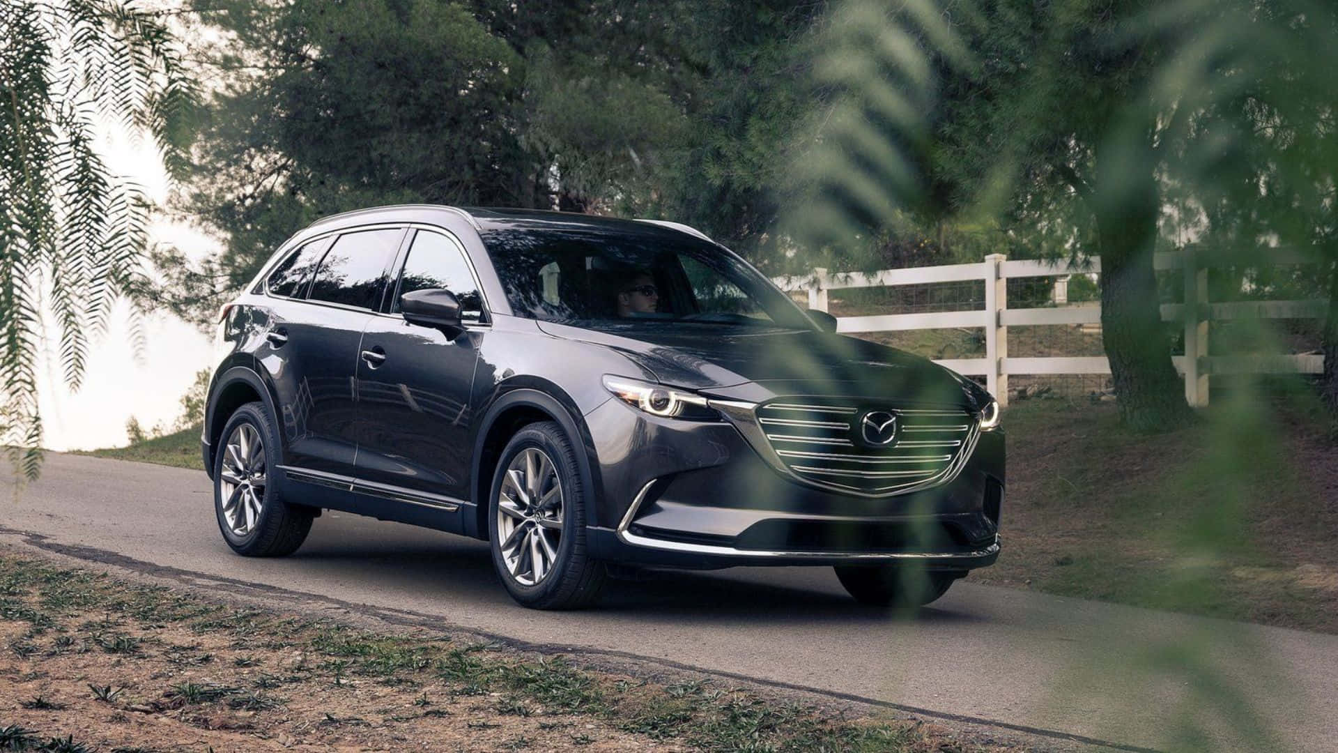 Mazda Cx-9 桌布 壁紙 — 一輛時尚的深灰色Mazda CX-9 SUV行駛在綠樹成蔭的鄉村道路上，背景是白色柵欄。