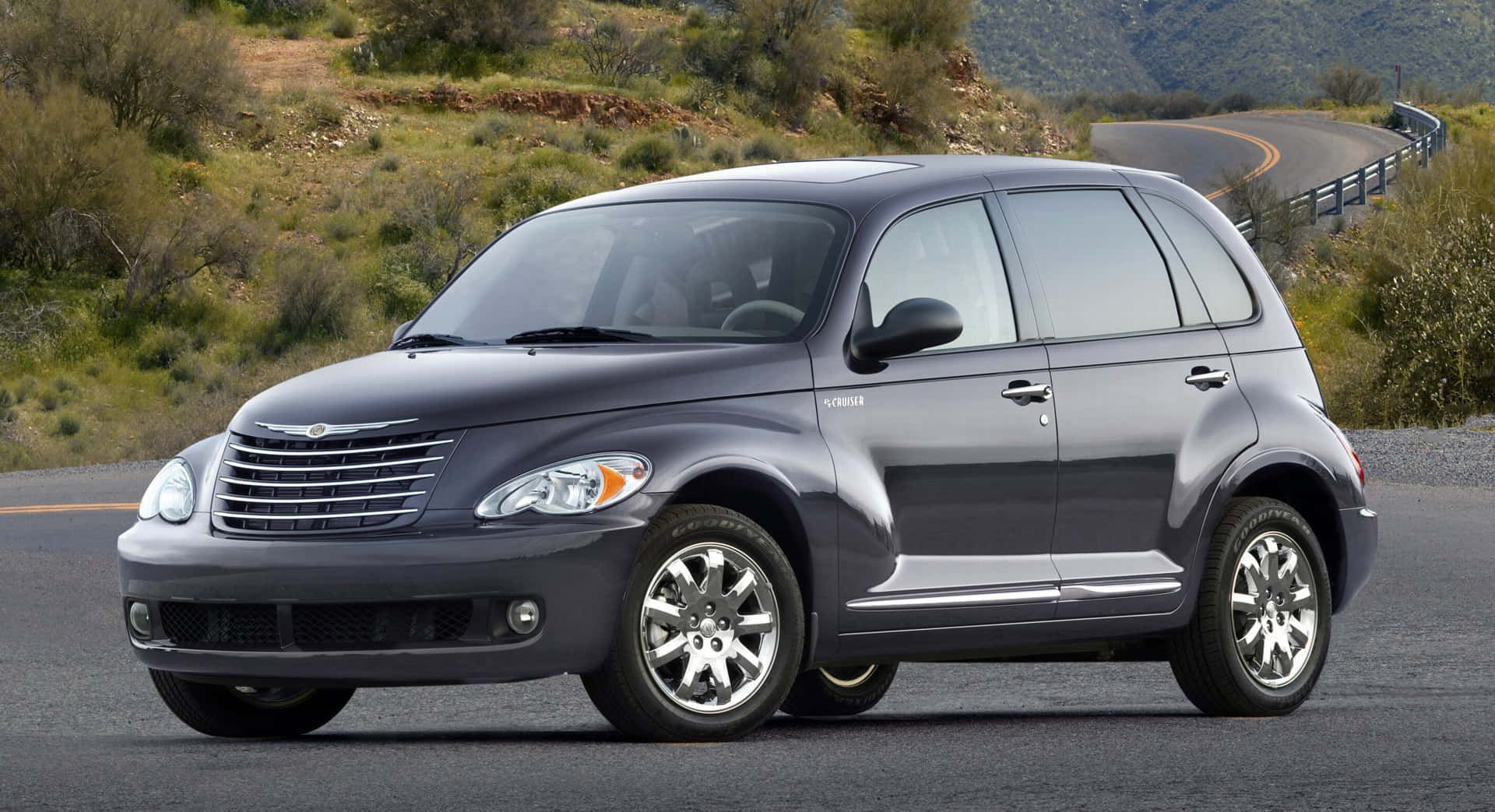 Chrysler Pt Cruiser 桌布 壁紙 — 一輛時尚的深灰色PT Cruiser在明亮的日光下行駛在蜿蜒的山路上，穿過乾燥的灌木叢和丘陵。