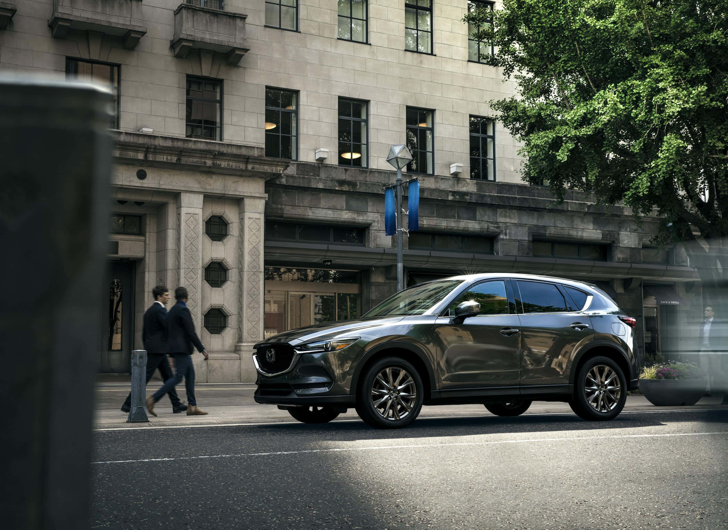 Mazda Cx-5 桌布 壁紙 — 一輛時尚的深灰色SUV駛過宏偉的石建築，兩名穿西裝的男子在明亮的日光下過馬路。