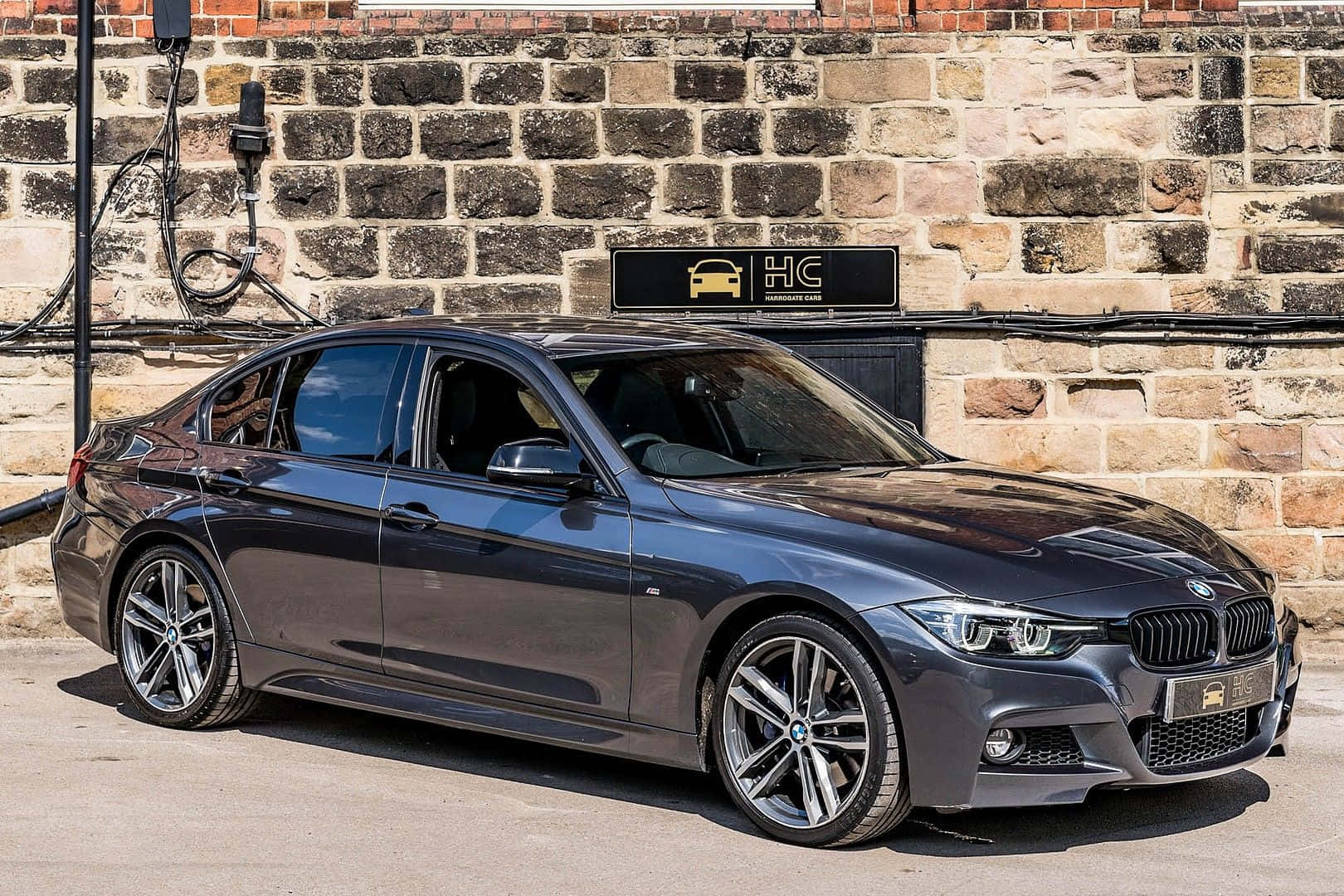 Bmw 3 Series 桌布 壁紙 — 一輛時尚的深灰色寶馬轎車在明亮的日光下停泊在充電站，背景是質樸的石牆。