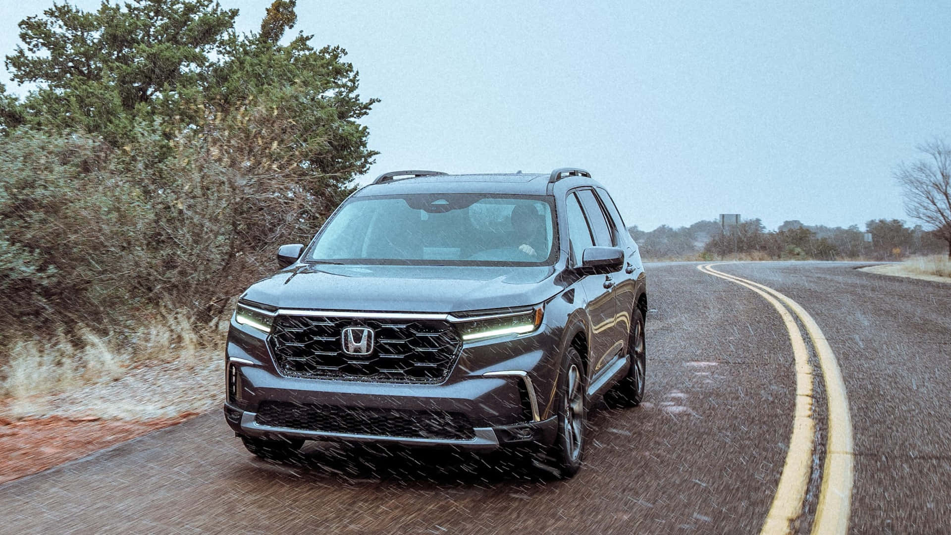 Honda Pilot 桌布 壁紙 — 一輛時尚的深灰色本田SUV在蒼白的天空下行駛在一條蜿蜒的沙漠公路上，兩旁是乾燥的灌木叢和遠處的樹木。