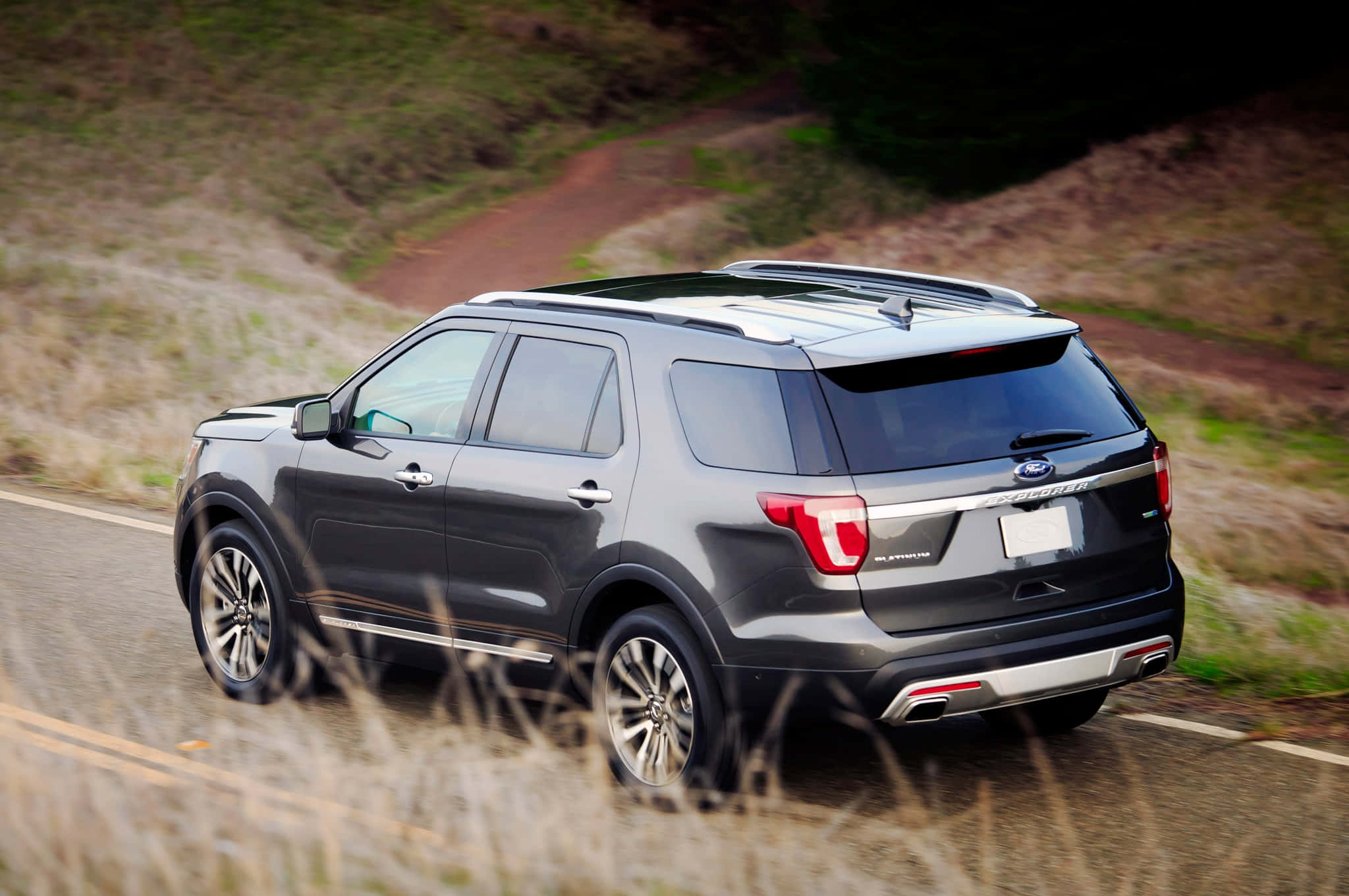 Ford Explorer 桌布 壁紙 — 一輛時尚的深灰色福特探險者SUV沿著蜿蜒的鄉村道路行駛，背景是模糊的草地和山丘。