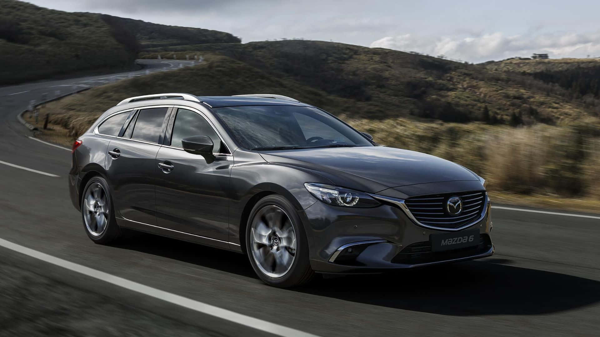 Mazda 6 桌布 壁紙 — 一輛時尚的深灰色萬事得6旅行車沿著蜿蜒的山路疾馳，在陰天背景下，模糊的綠色山坡一閃而過。