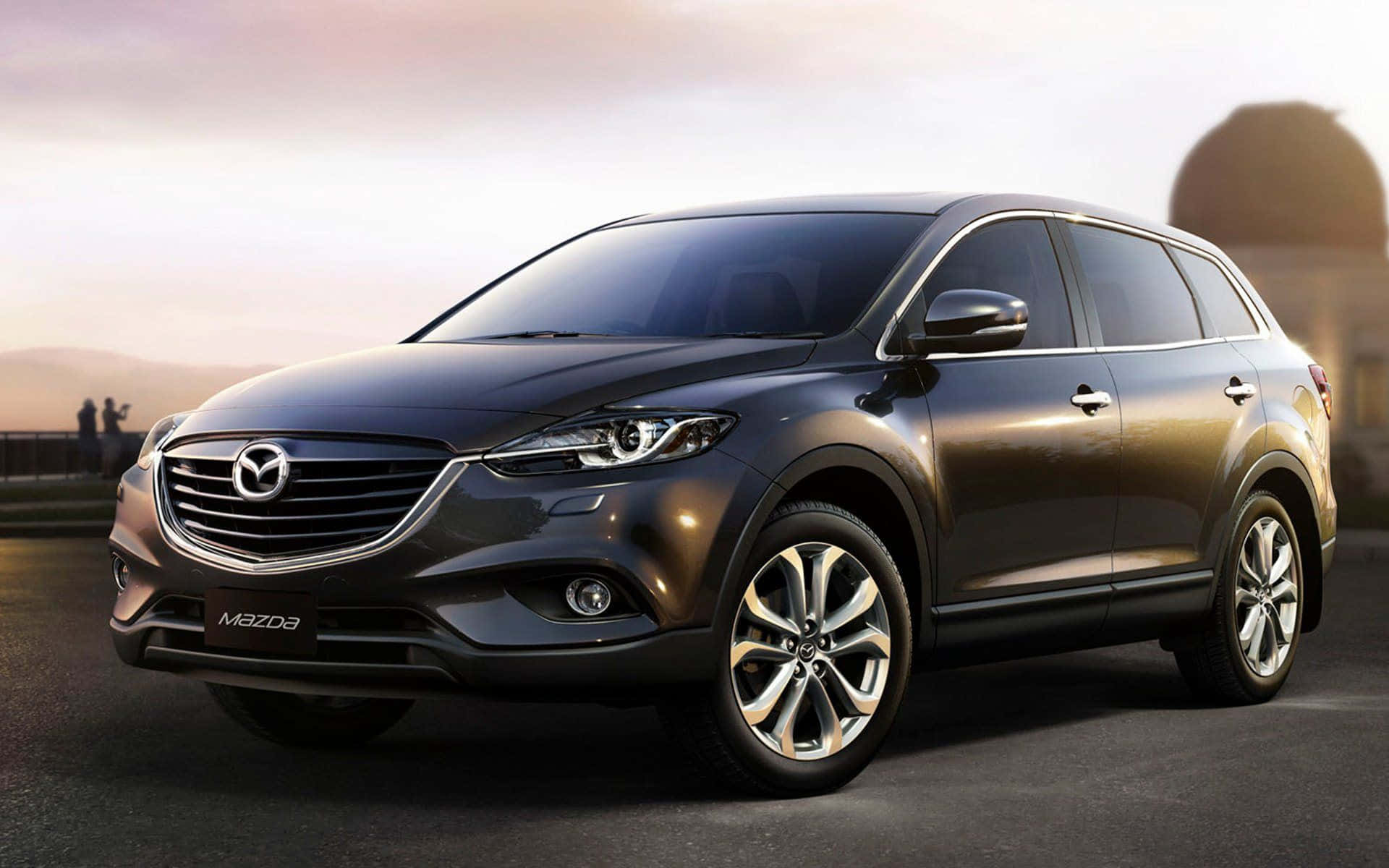 Mazda Cx-9 桌布 壁紙 — 一輛時尚的深灰色萬事得CX-9 SUV在溫暖的日落光線下，在鋪砌的道路上閃閃發光，遠方有山丘和剪影人物。