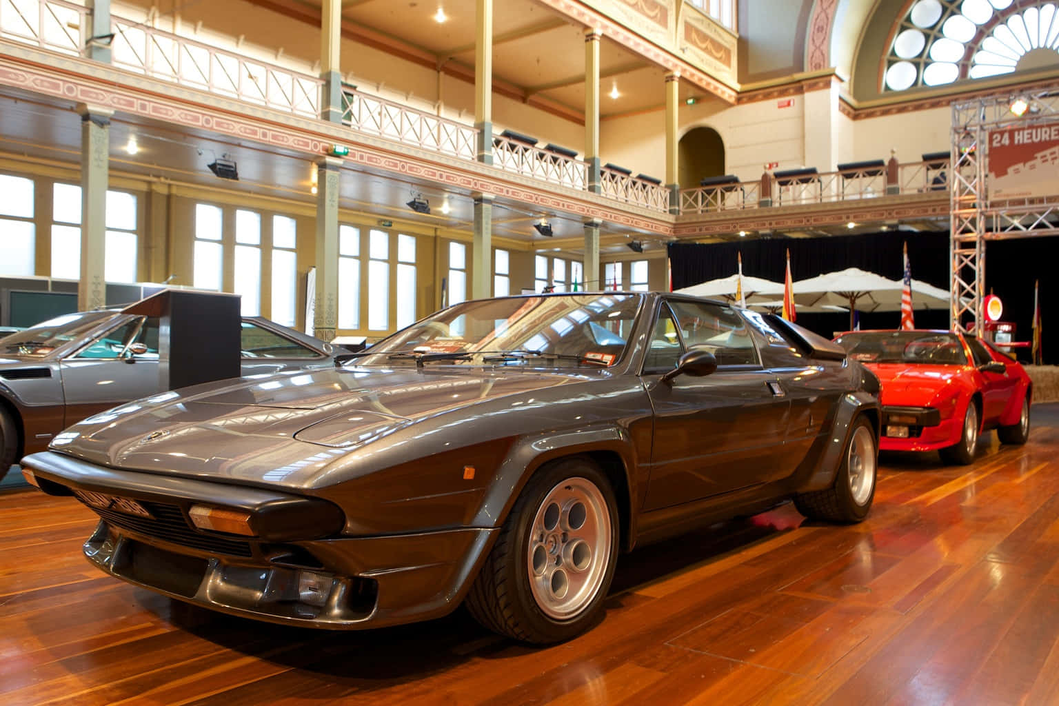 Lamborghini Silhouette 桌布 壁紙 — 一輛時尚的深灰色跑車在明亮的燈光下，在宏偉的多層展覽廳內的拋光木地板上閃閃發光。