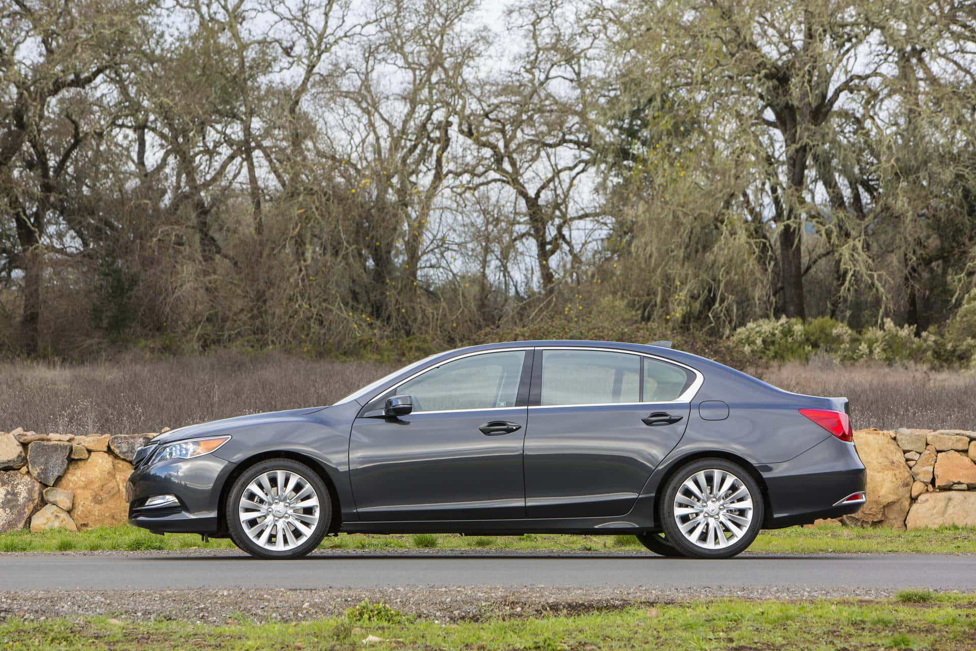 Acura Rlx 桌布 壁紙 — 一輛時尚的深灰色轎車停泊在碎石路上，旁邊是石牆，背景有光禿禿的樹木。