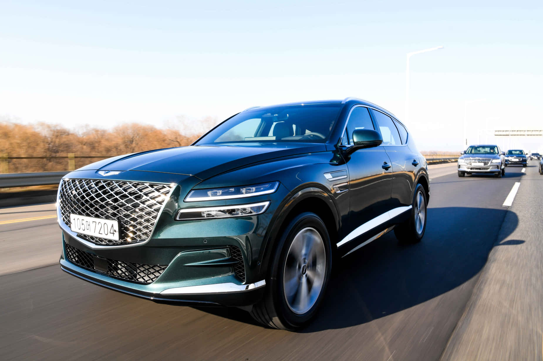 Genesis Gv80 桌布 壁紙 — 一輛時尚的深綠色 Genesis GV80 SUV 在陽光明媚的高速公路上行駛，路邊種滿了秋天的樹木。
