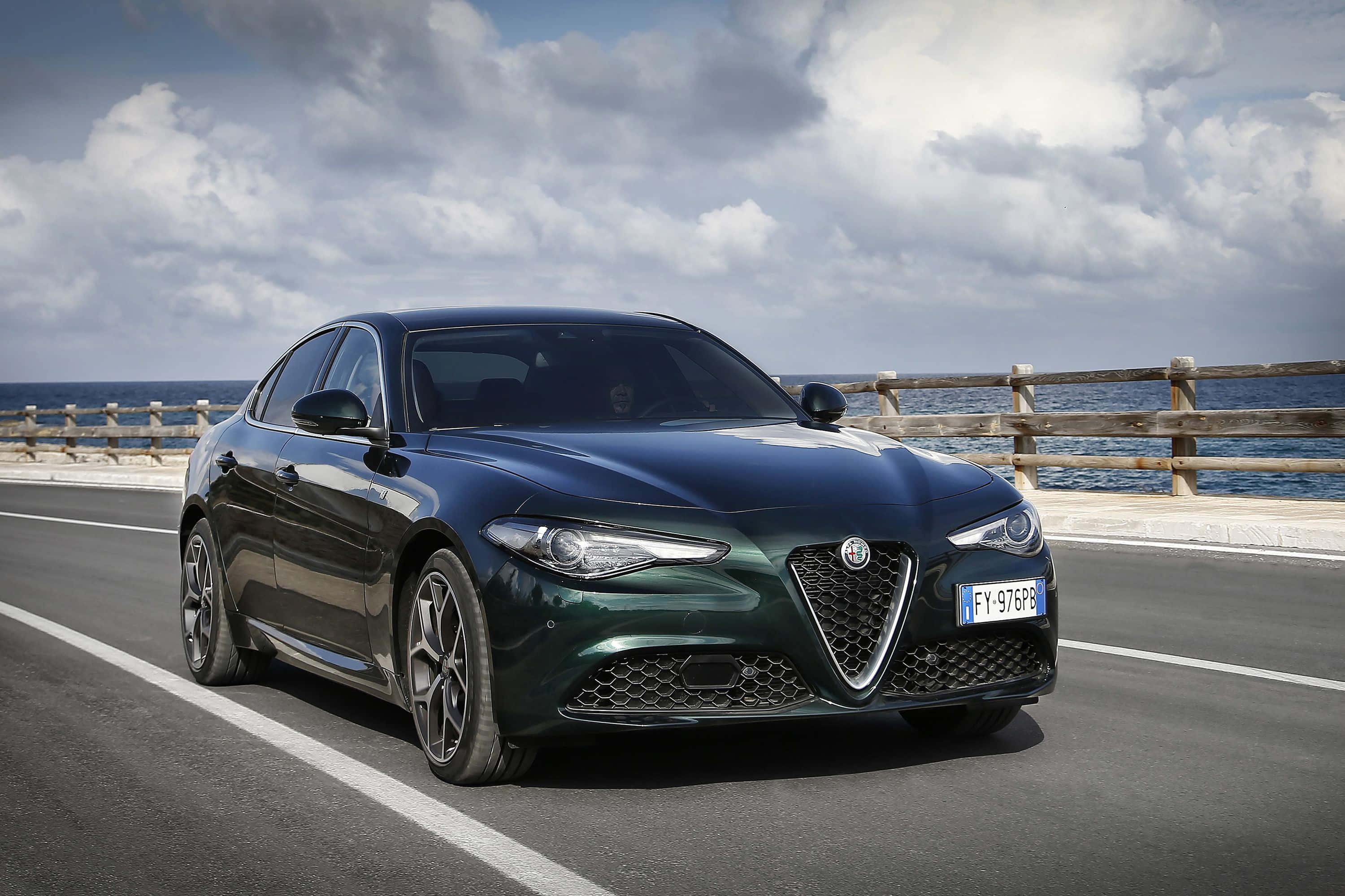 Alfa Romeo Stelvio 桌布 壁紙 — 一輛時尚的深綠色Alfa Romeo房車沿著海岸道路行駛，背景是海洋和多雲的天空。