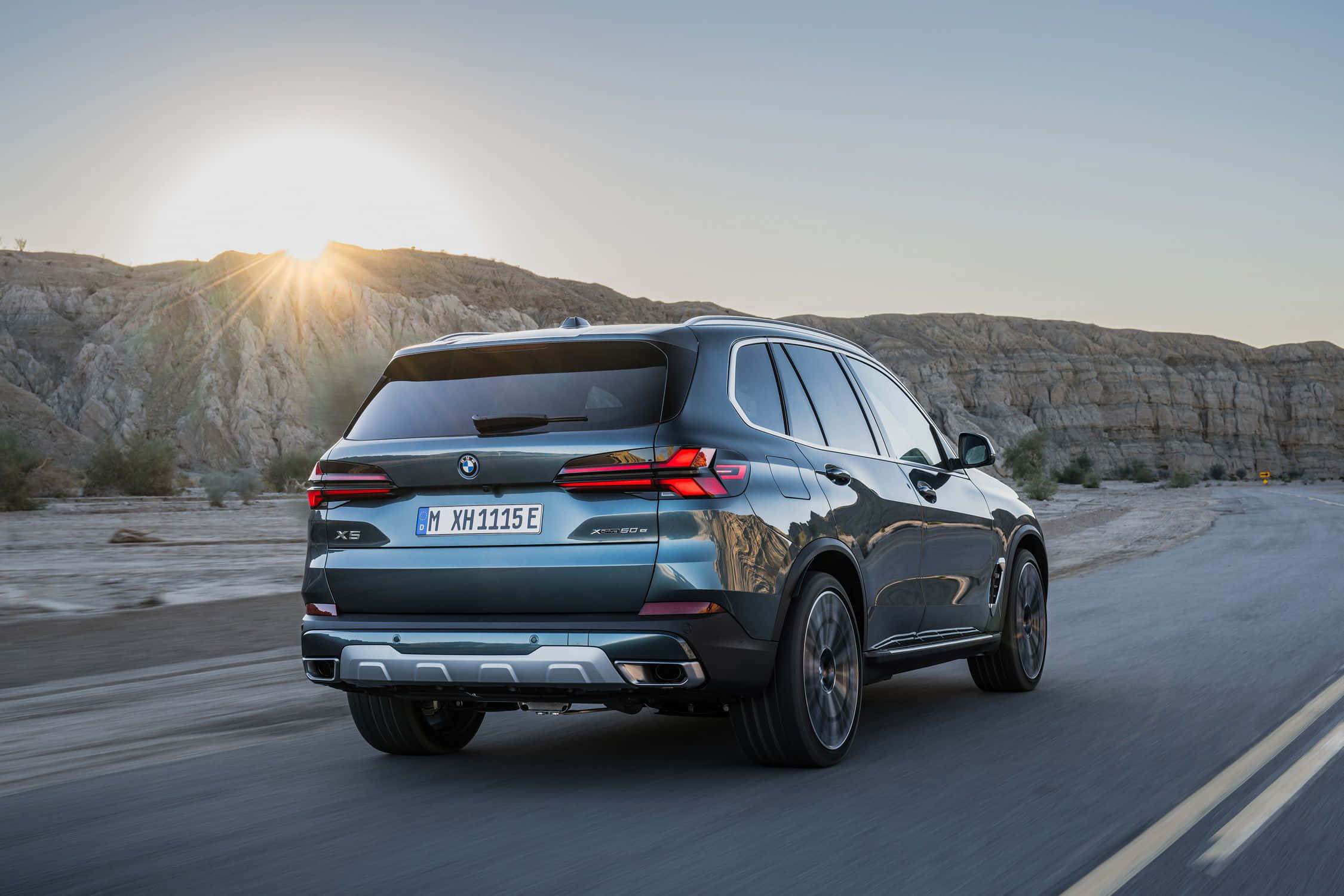 Bmw X5 桌布 壁紙 — 一輛時尚的深綠色BMW X5 SUV在沙漠公路上行駛，夕陽落在岩石山丘後方，灑下金色光芒。