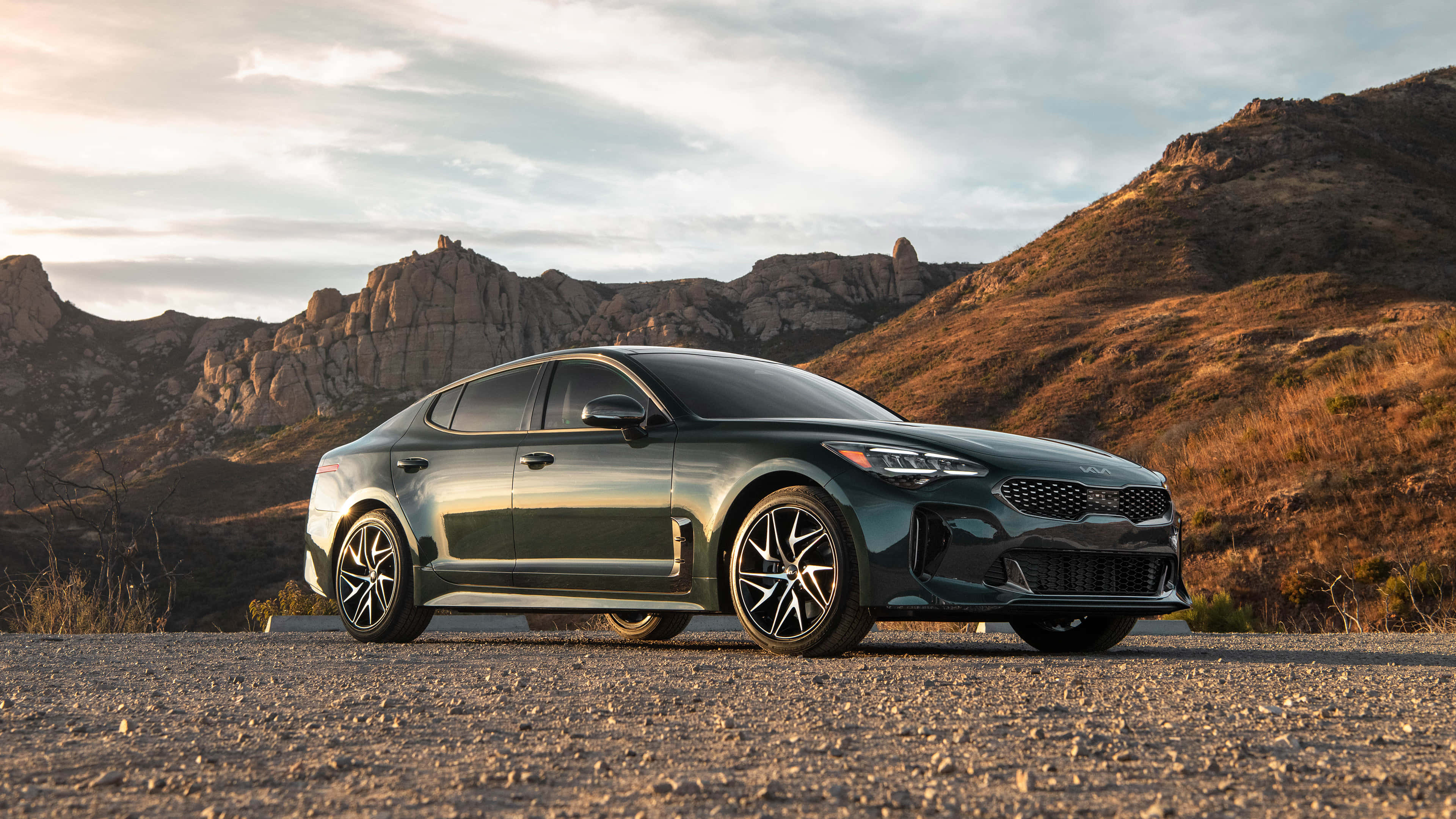 Kia Stinger 桌布 壁紙 — 一輛時尚的深綠色Kia轎車停在碎石路上，背景是崎嶇的山脈和溫暖的金色光芒。