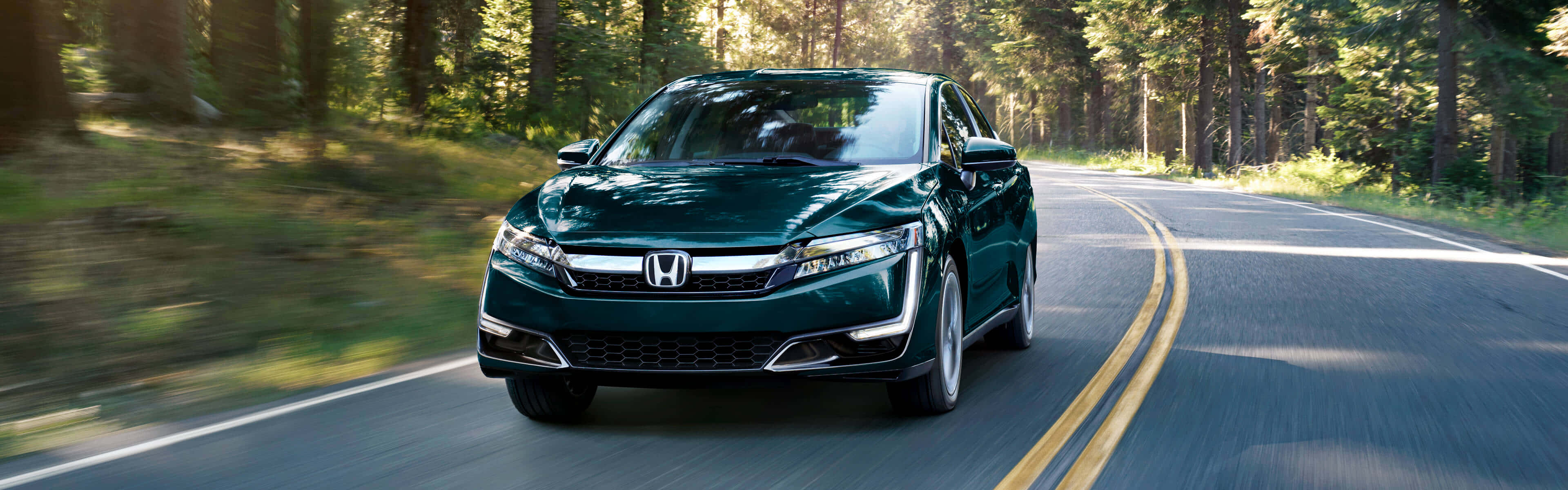 Honda Clarity 桌布 壁紙 — 一輛時尚的深綠色本田房車在蜿蜒的森林道路上飛馳，樹木模糊，陽光透過樹冠灑落。